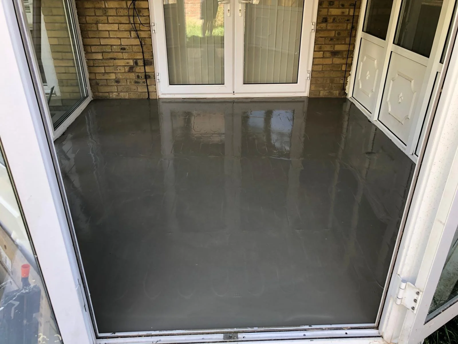 A freshly poured wet concrete floor in a sunroom enclosed with glass doors and brick walls.