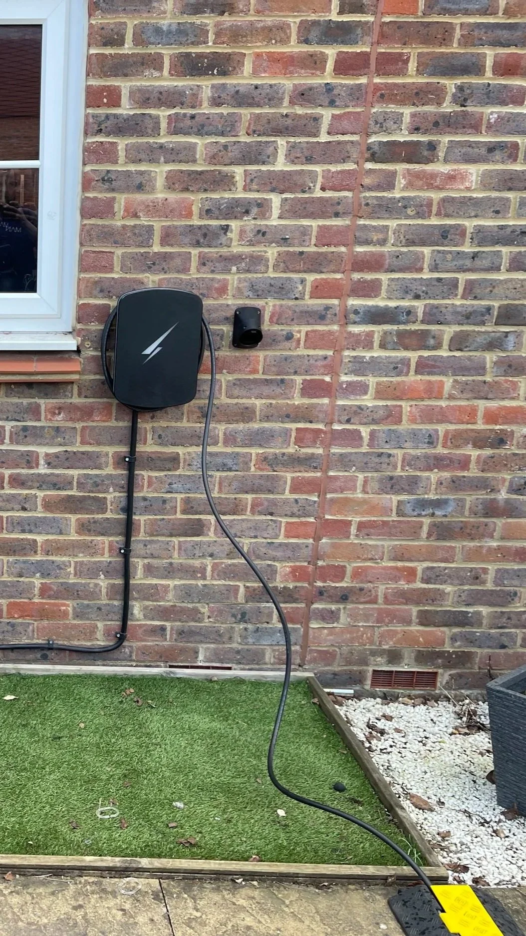 External view of a brick house wall with a green artificial grass patch and a charging station for an electric vehicle, with cables connected.