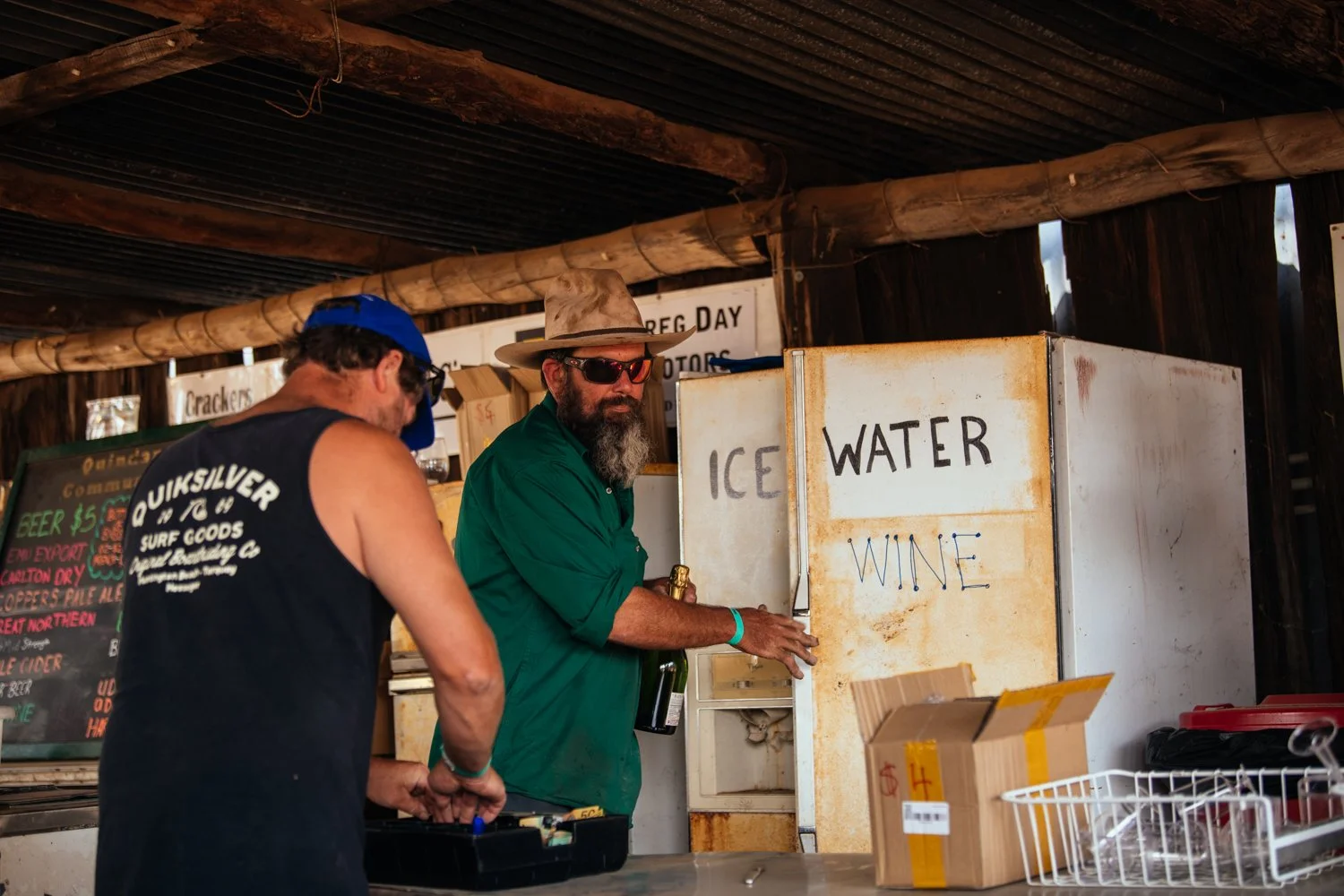Two men at a rustic outdoor refreshment stand, one in a black tank top and blue cap, the other in a green shirt, wearing a hat and sunglasses, near a rusted white fridge labeled 'ICE WATER' and 'WINE,' with cardboard boxes and a chalkboard menu in the background.