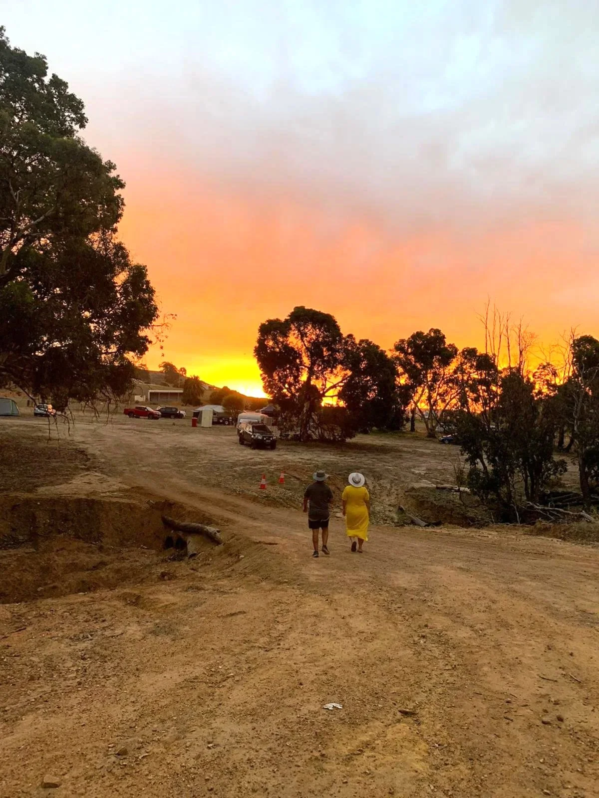 A dirt road with two people walking away from the camera during a sunset. The sky is filled with orange and pink clouds, and there are trees and parked cars in the background.