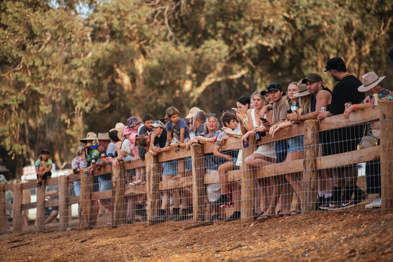 A group of people, including children and adults, leaning on a wooden fence, watching something in the distance at an outdoor event with large trees in the background.