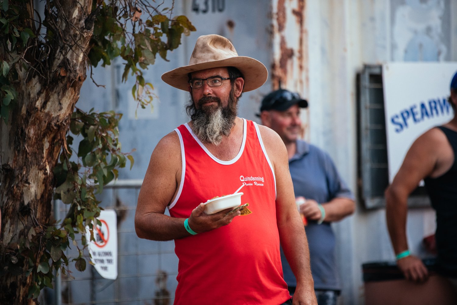 A man with a beard, glasses, and a tan hat wearing a red tank top, holding a bowl of food at an outdoor event. Other people are in the background.