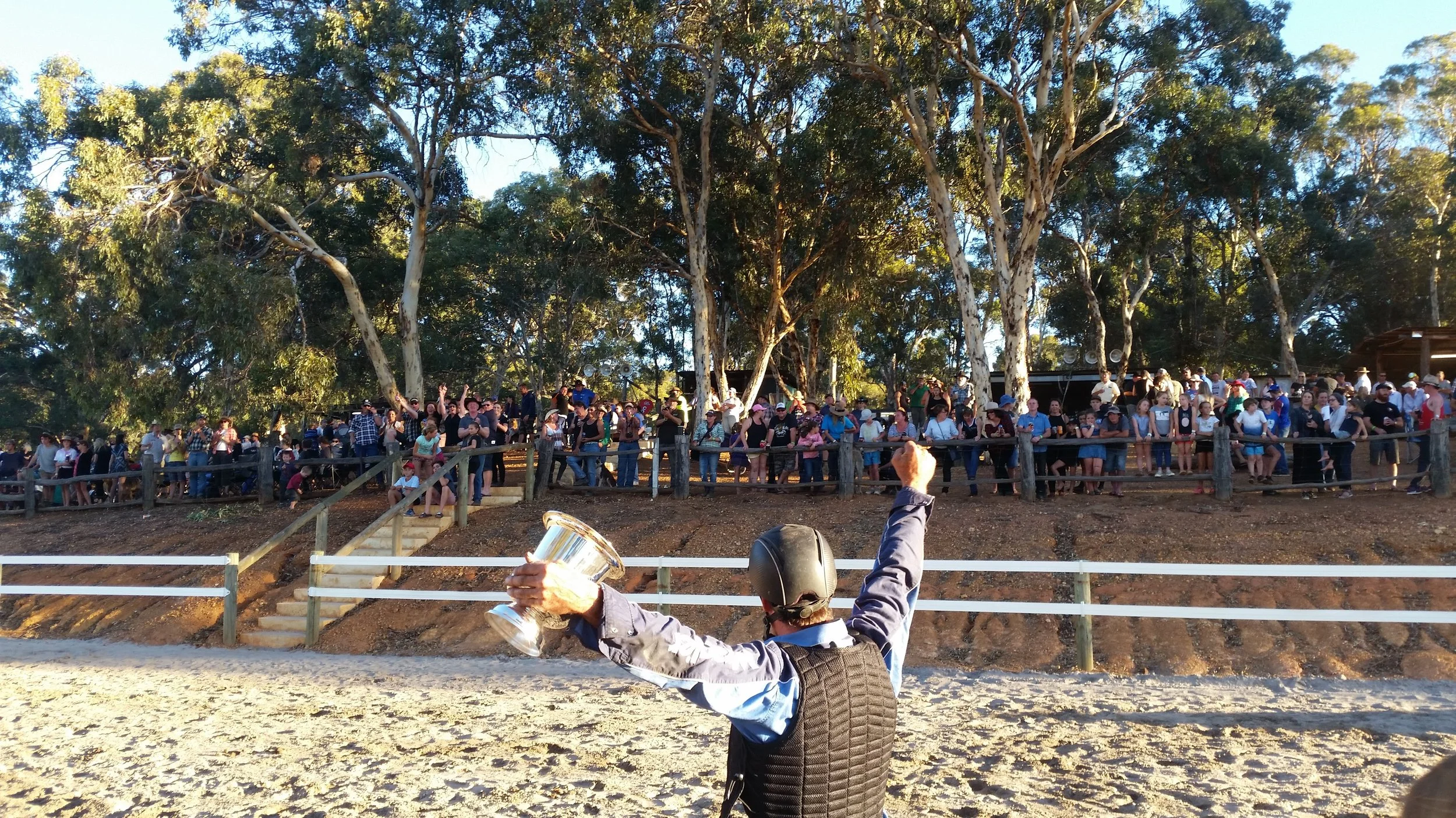 A person wearing a helmet and vest is holding a trophy in one hand and raising a fist in the air while facing a large crowd behind a wooden fence at an outdoor event surrounded by tall trees.