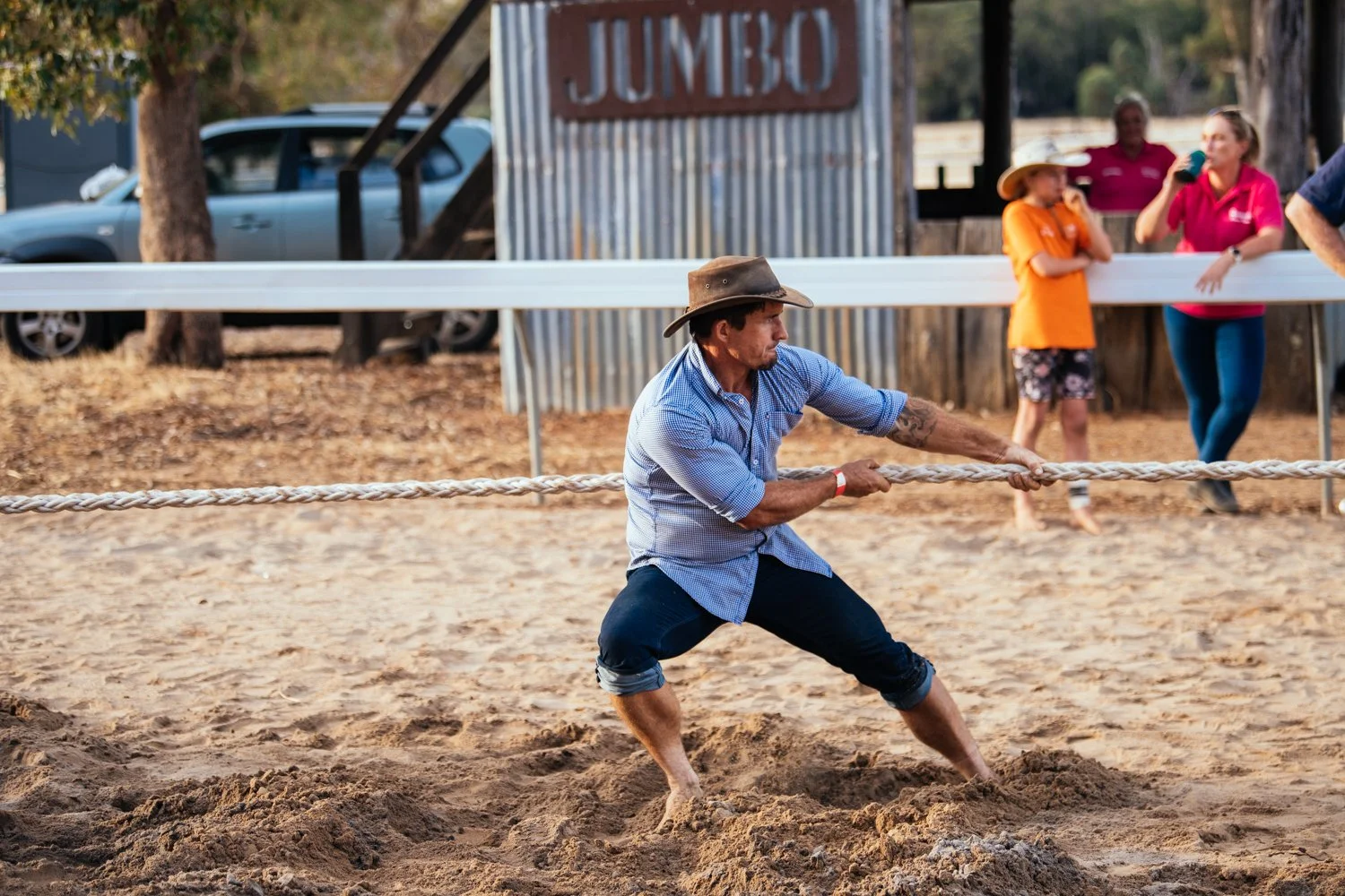 A man in a blue checkered shirt and rolled-up jeans, barefoot, participating in a tug-of-war game at an outdoor event, with spectators in the background.