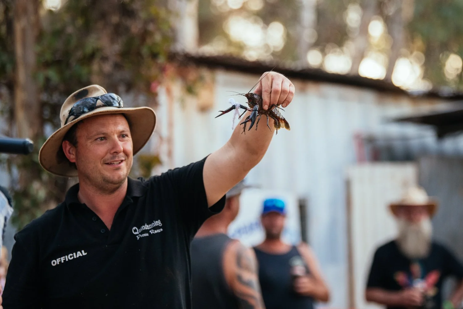 A man wearing a wide-brimmed hat and a black shirt labeled "Official" is holding a large crawfish or lobster in one hand, raised high. In the background, a group of people are gathered outdoors, some in hats, with a relaxed atmosphere.