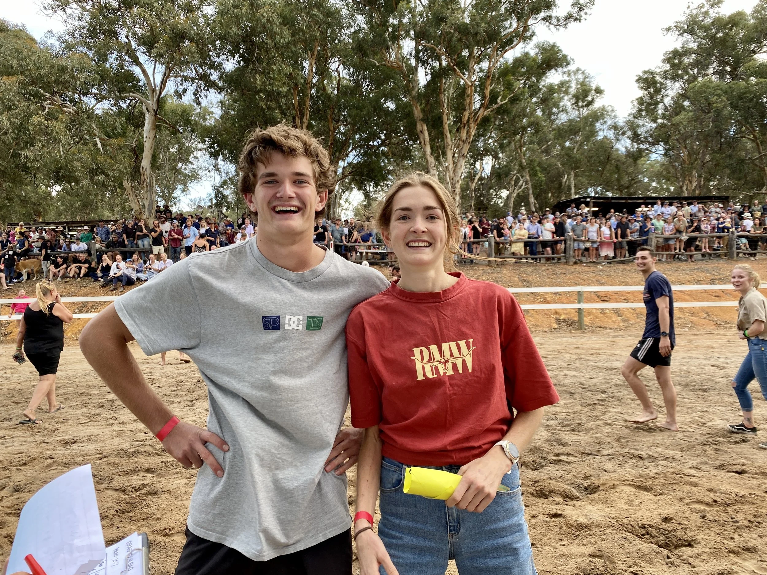 Two young adults, a man and a woman, smiling and standing on a sandy outdoor area with a crowd of people behind a fence and tall trees in the background.
