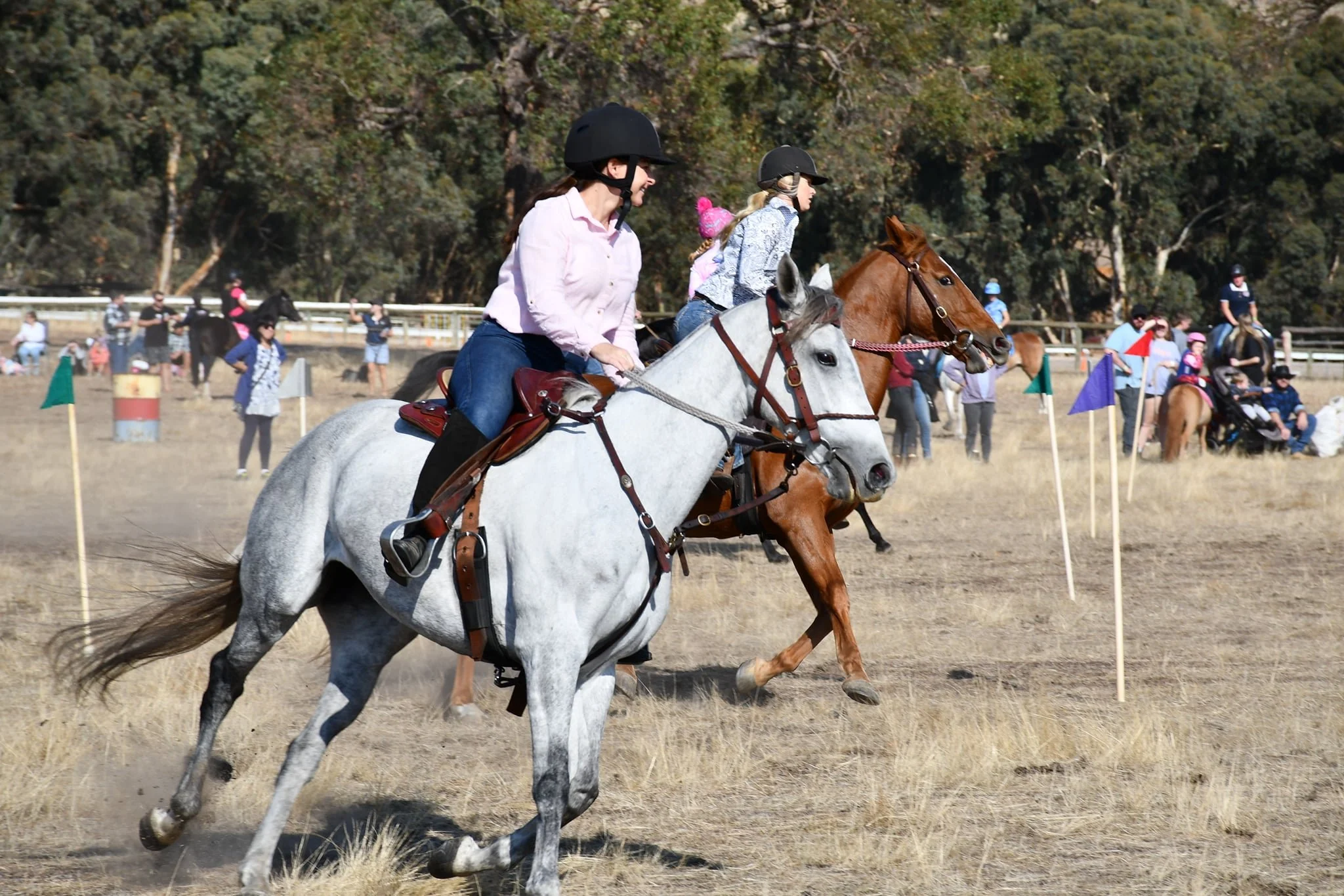 Two women riding horses in an outdoor field with spectators and trees in the background
