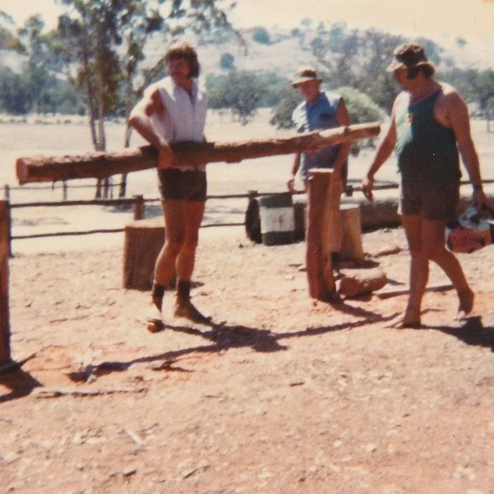 Three women outdoors lifting a large wooden log between two wooden posts.