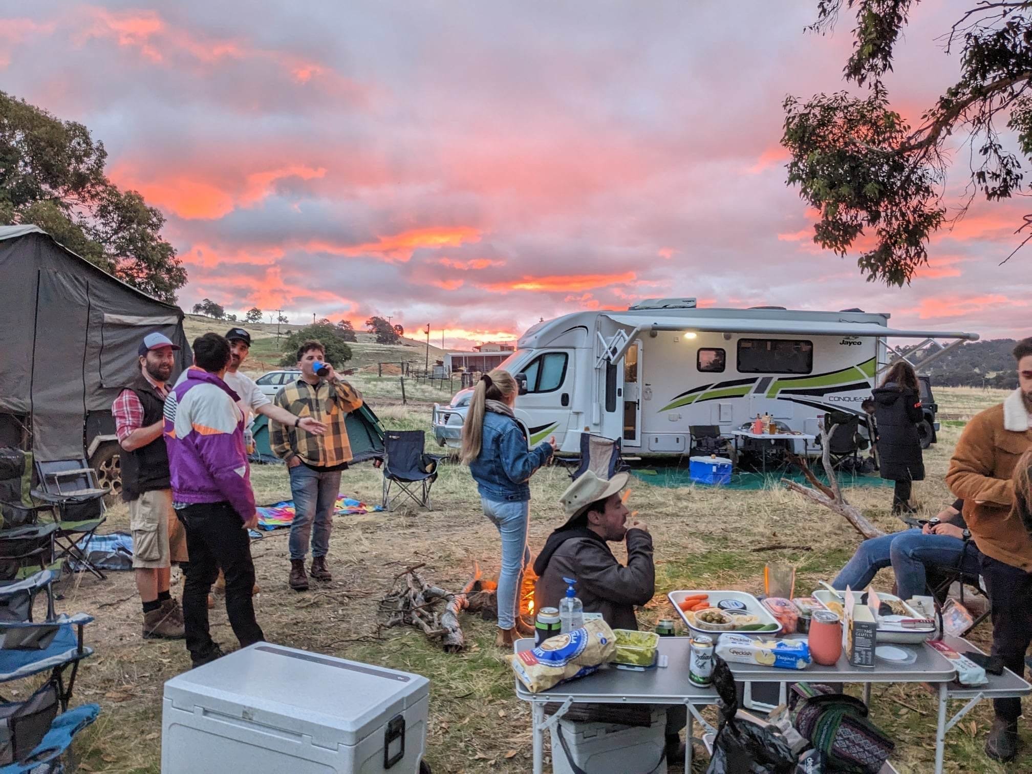 Group of people gathered outdoors around a campground with a RV, picnic table with food, and chairs during sunset, in a rural setting.