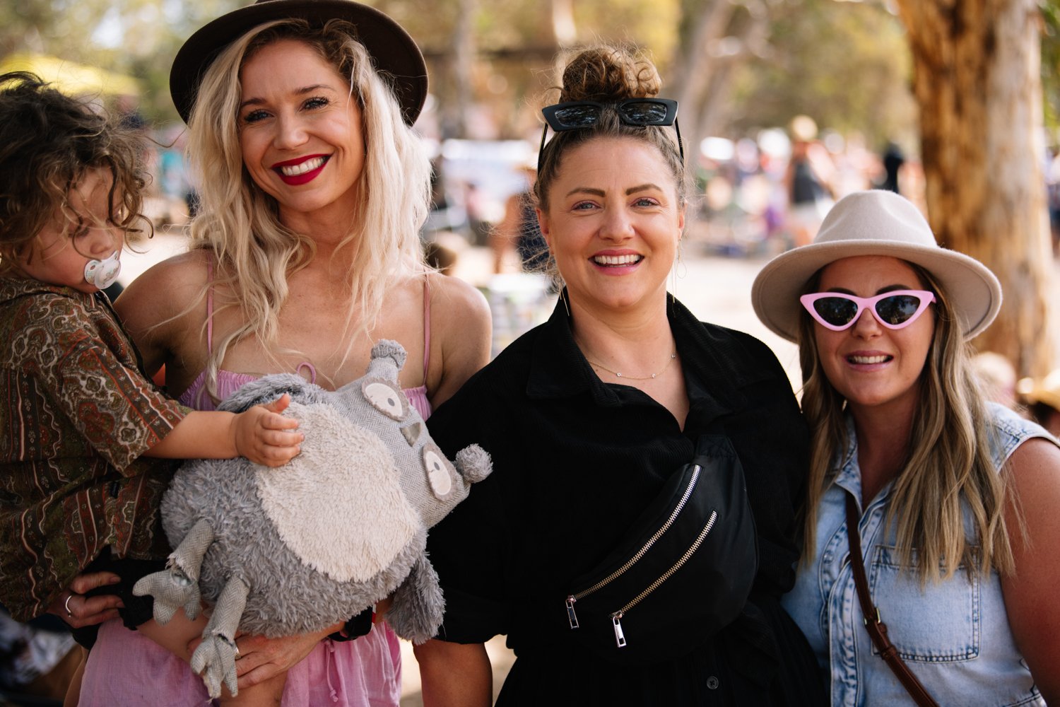 Three women smiling outdoors at a park, with trees and people in the background, wearing casual summer clothing and fashionable accessories.