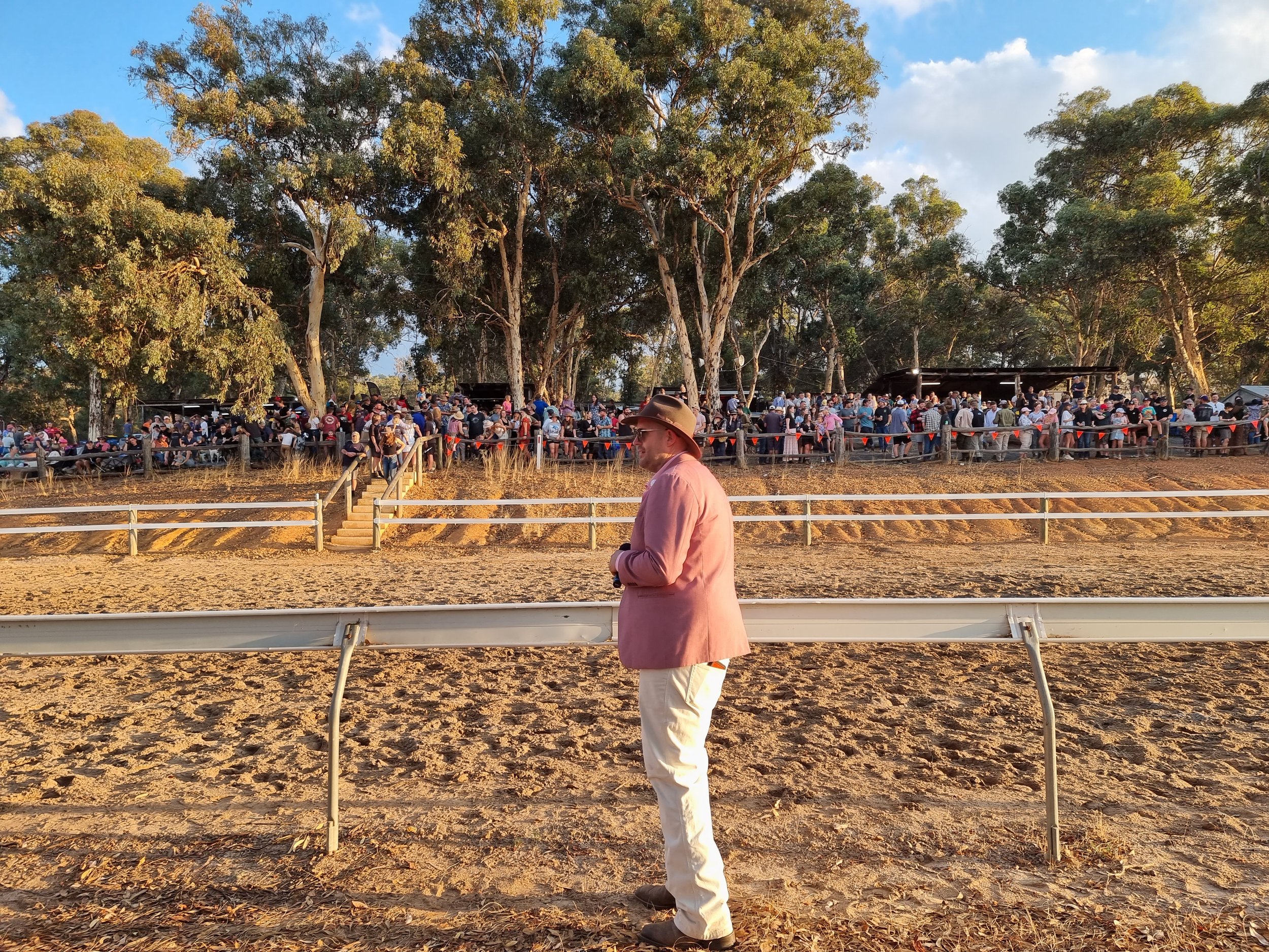 A man in a pink jacket and beige pants standing in front of a racetrack with a large crowd of people gathered in the background under trees, in the late afternoon.