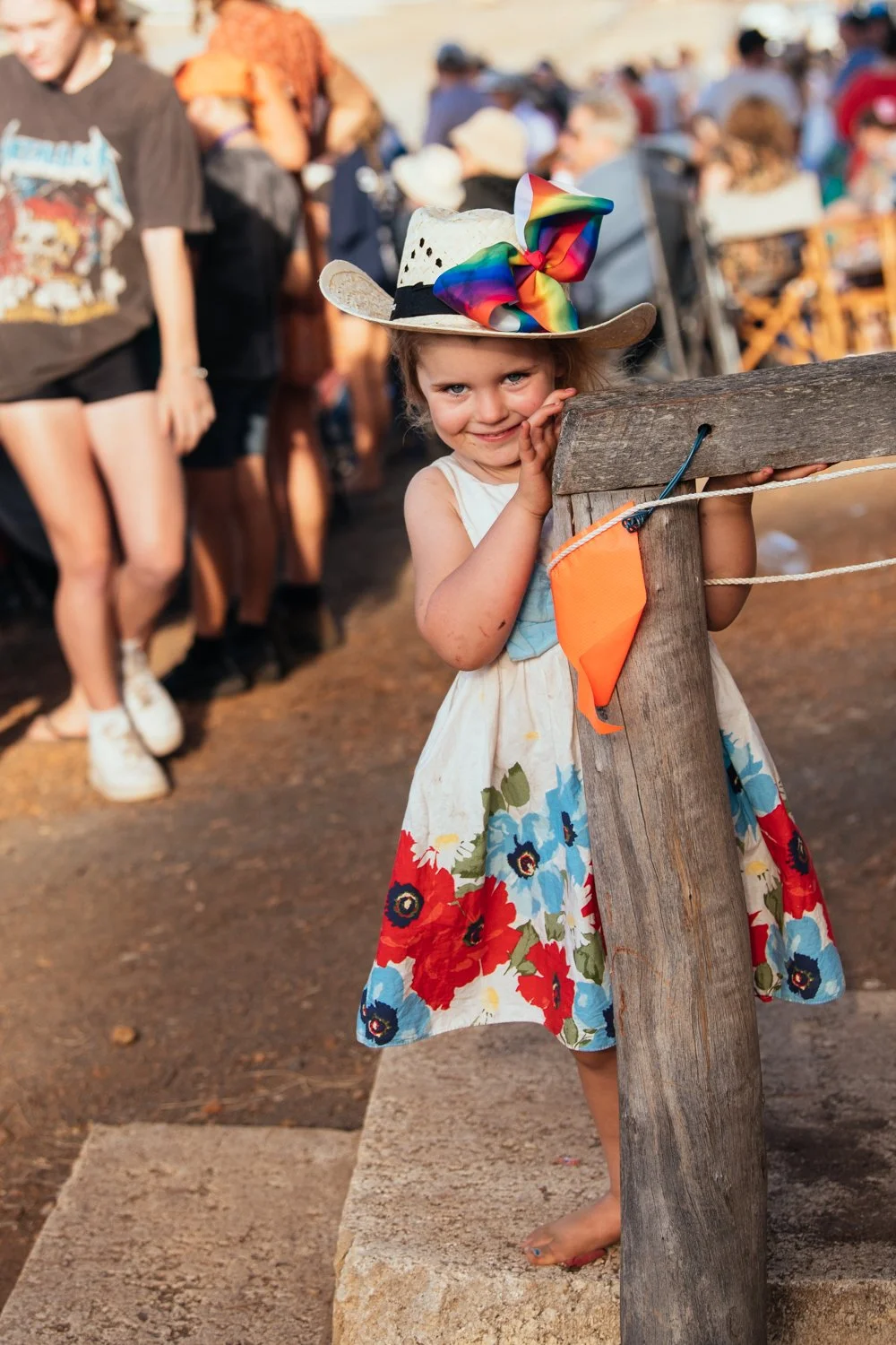 A young girl in a floral dress with red, blue, and white flowers, wearing a cowboy hat with a rainbow-colored bow, leaning on a wooden post at an outdoor event with a crowd of people in the background.