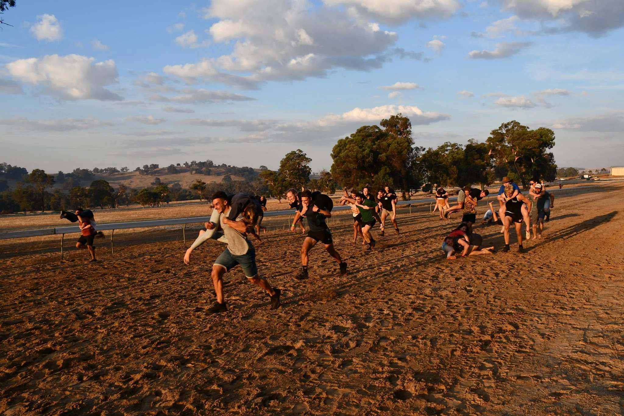 People participating in a piggyback race on a dirt track during sunset, with trees and hills in the background.