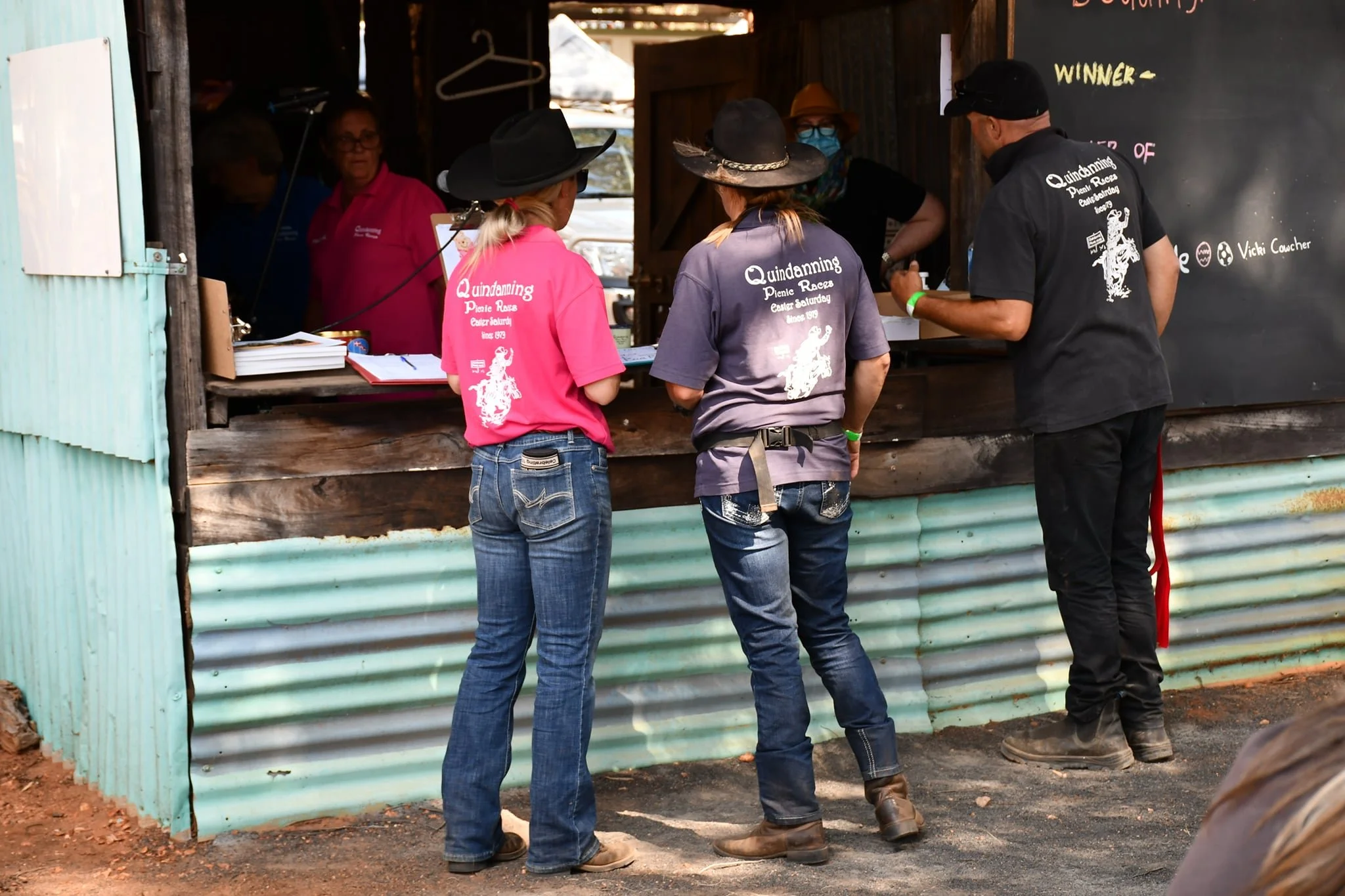 Three women and one man at the ticket counter of the Quindanning Picnic Races, an outdoor event with a rustic, informal setting. The women are wearing pink and purple shirts, and the man is in black. They are engaged in conversation with the person behind the counter inside a wooden booth.