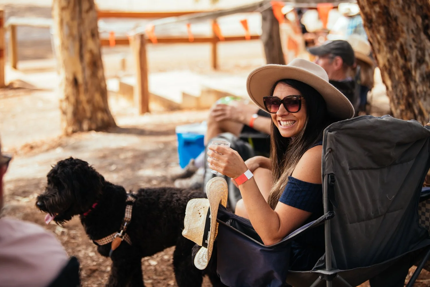 Woman wearing sunglasses and a wide-brimmed hat sitting on a camping chair, smiling with a drink in her hand, next to a black dog with a red collar outdoors.