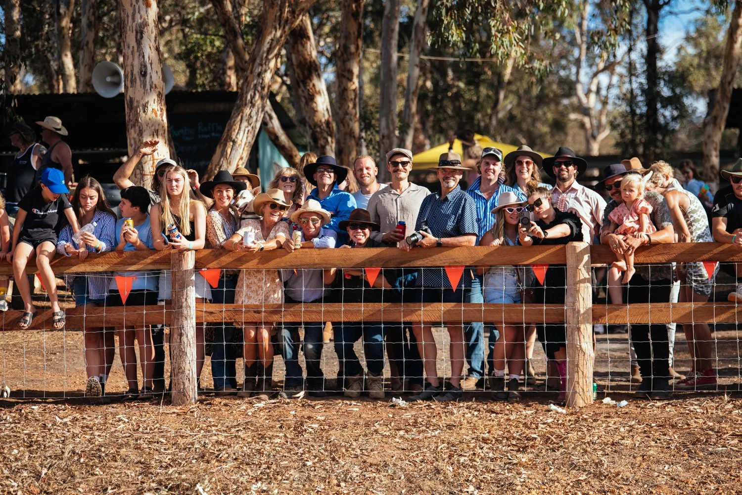 Group of people gathered behind a wooden fence outdoors, some wearing hats and sunglasses, with trees and blue sky in the background