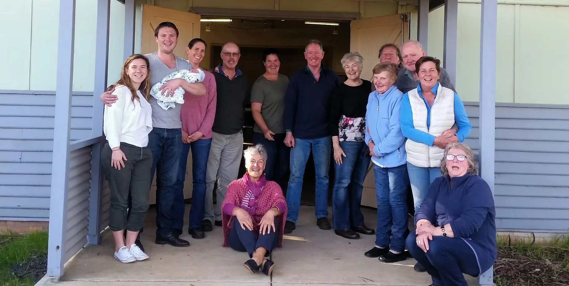 Group of people, including children and older adults, standing and sitting on the porch of a house, smiling for the photo.