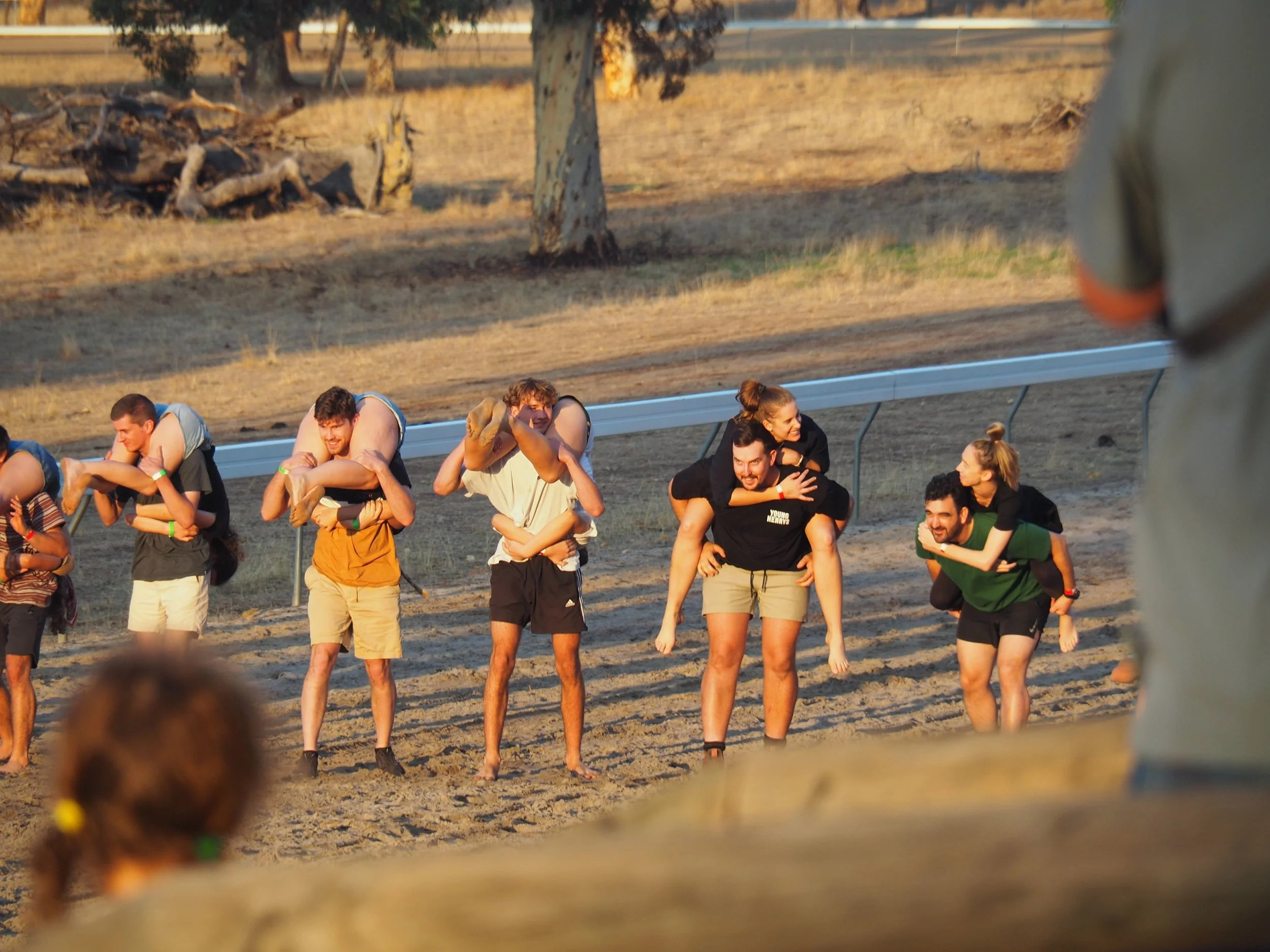 Group of people in a piggyback race on a dirt field during a sunny day.