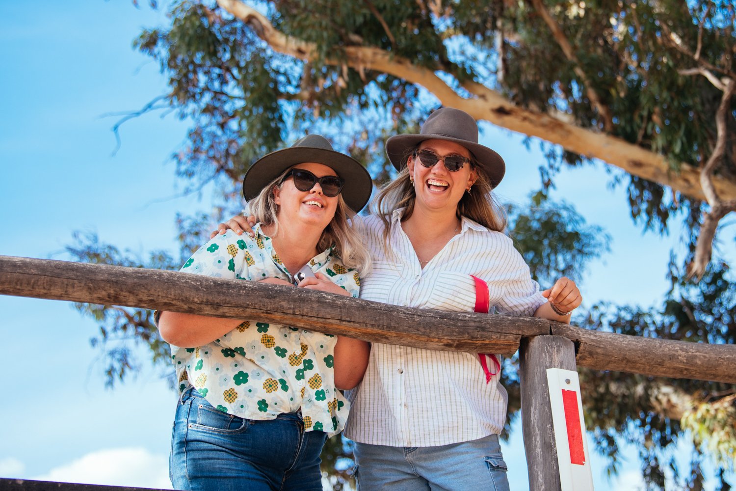 Two middle-aged women wearing hats and sunglasses, smiling and laughing while leaning on a wooden railing outdoors with trees and blue sky in the background.