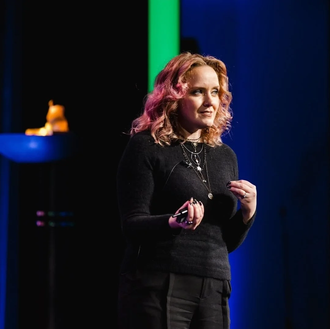 A woman with wavy, blonde hair, wearing a black long-sleeve top and necklaces, giving a presentation on stage with a dark background and pink, green, and blue lighting.