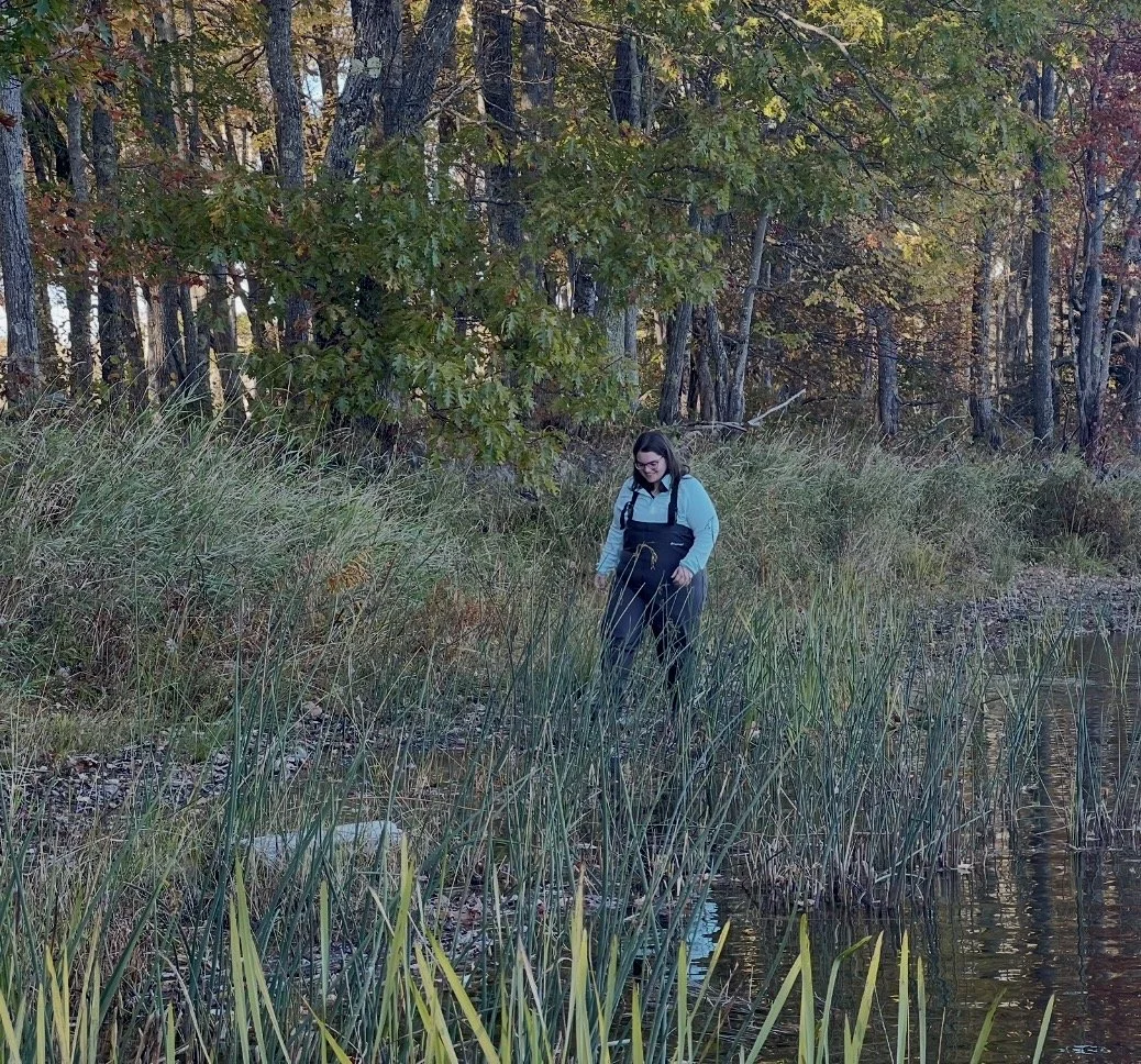 A woman in outdoor gear standing near water surrounded by tall grasses and trees, during autumn.