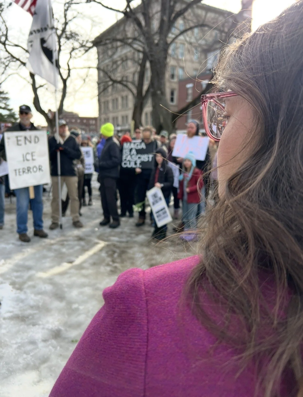 Crowd of protesters holding signs at a rally, with a woman in pink glasses and a magenta jacket in the foreground