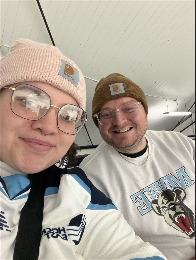 Two people wearing winter hats and glasses, smiling for a selfie indoors. The woman on the left has a pink beanie, large glasses, and a white jacket. The man on the right has a brown beanie, glasses, and a white sports jersey with a roaring lion grap