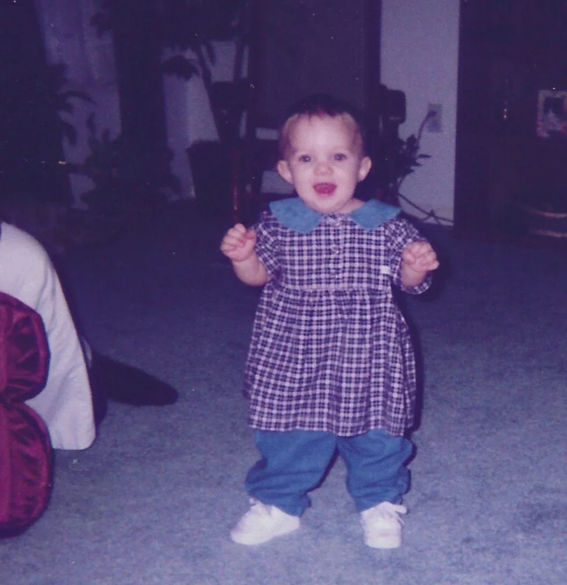 A young toddler standing indoors with arms slightly bent and fists clenched, smiling, wearing a checkered dress with a blue collar, blue pants, and white shoes.