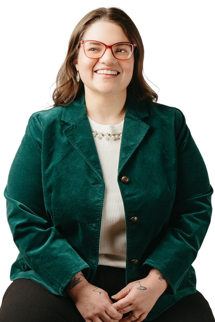 A young woman with shoulder-length brown hair, wearing red glasses, a green blazer, a white top with pearl detail, and earrings, smiling while sitting with her hands clasped in her lap.