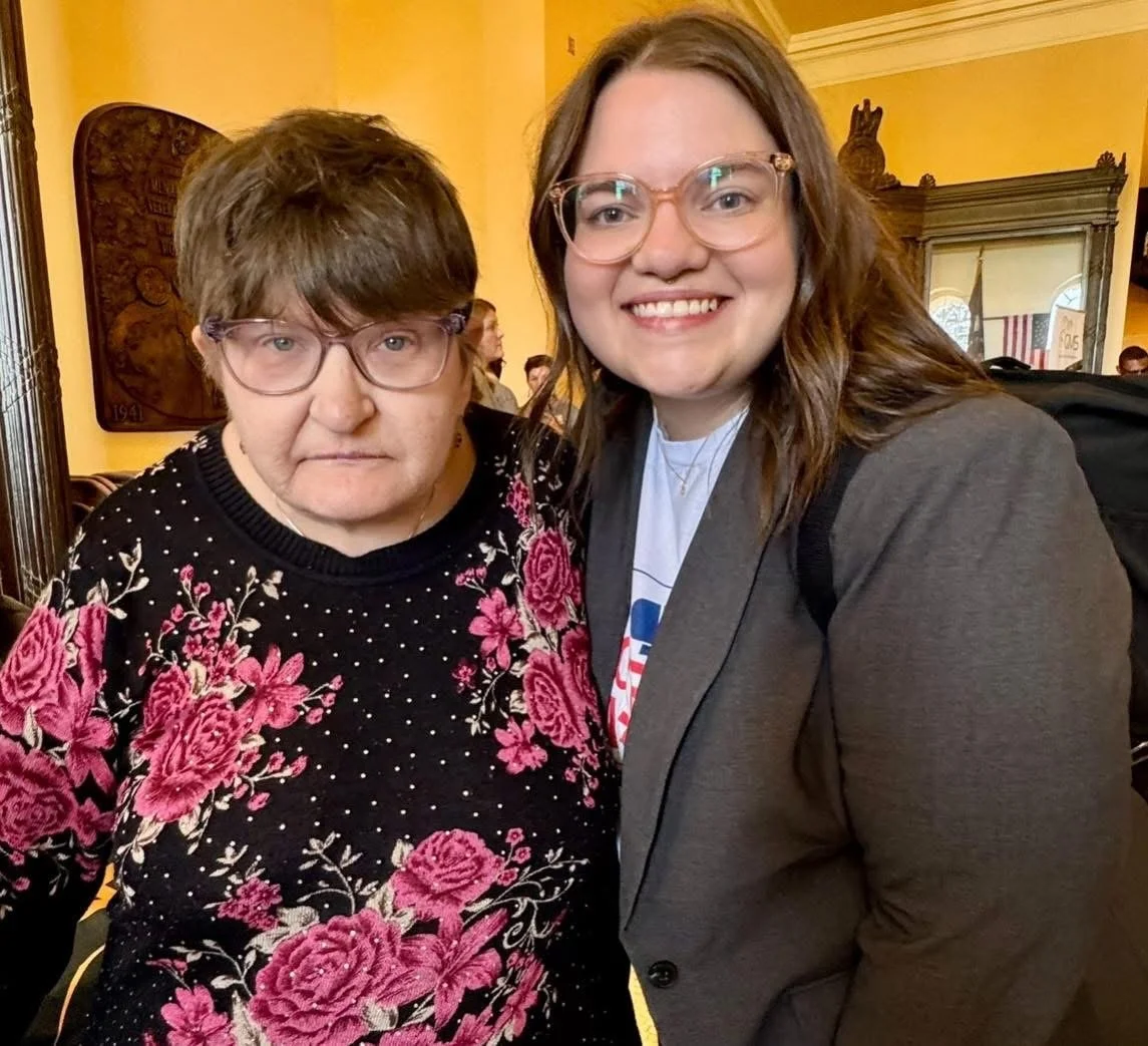 Two women smiling for the camera indoors, one with glasses and a floral sweater, and the other with glasses and a blazer, in a room with yellow walls and framed artwork.