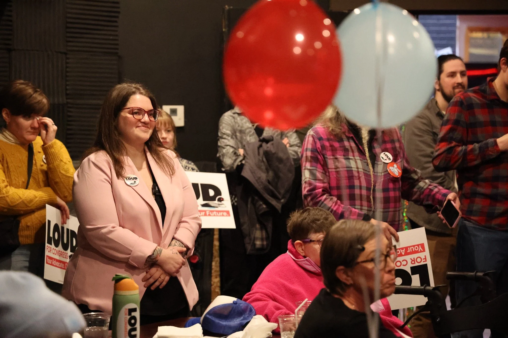 People gathered at a political rally, some holding signs that say 'LOUD FOR CONGRESS Your Advocate' and wearing campaign buttons.