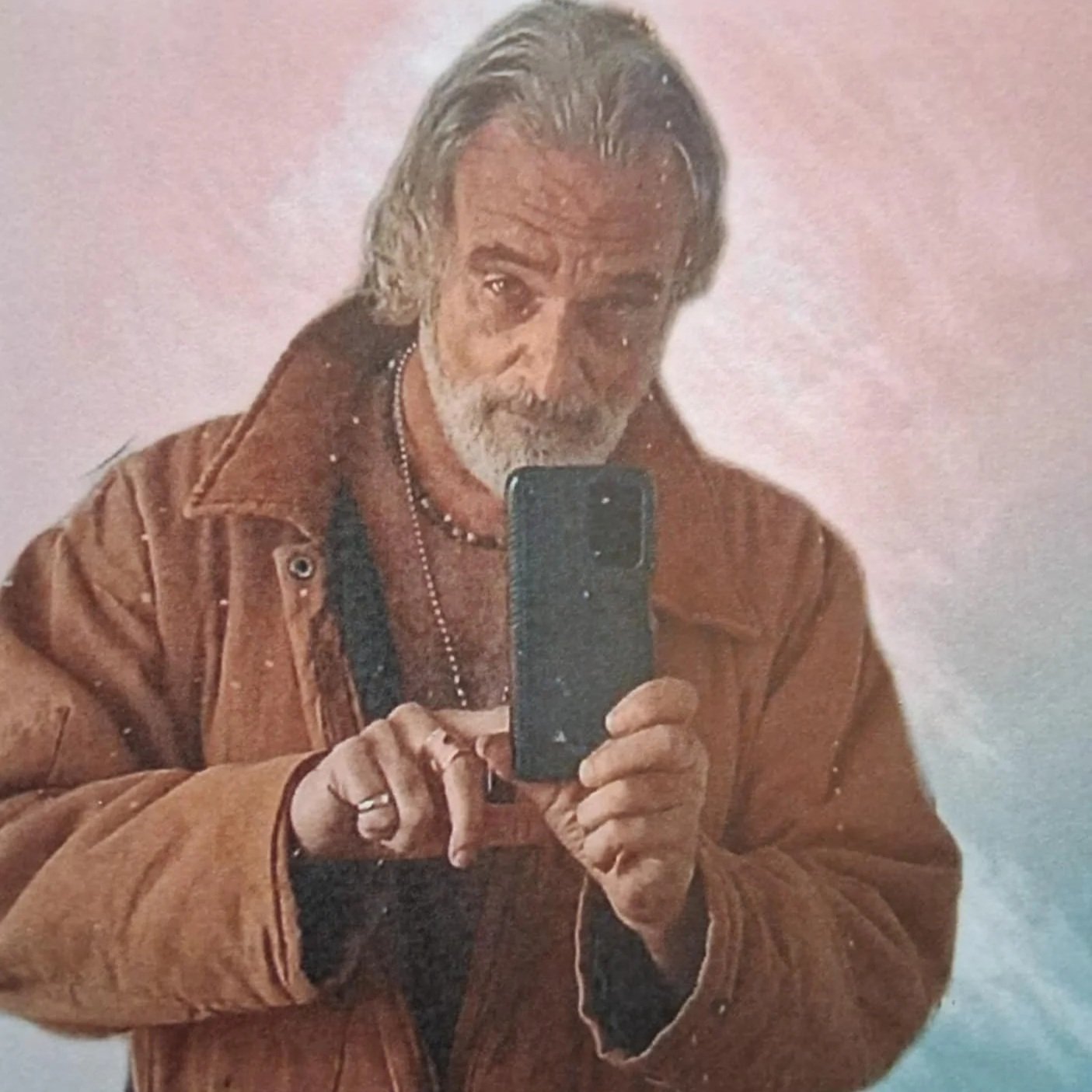 A man with gray hair and a beard taking a photo of himself in a mirror with a smartphone. He is wearing a brown jacket, black shirt, and a necklace.