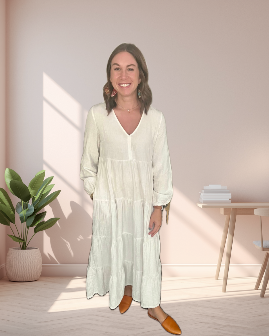 Valerie Marston, business owner, standing in a room with natural light, wearing a long white dress and brown shoes, with a potted plant and a small table with stacked books in the background.