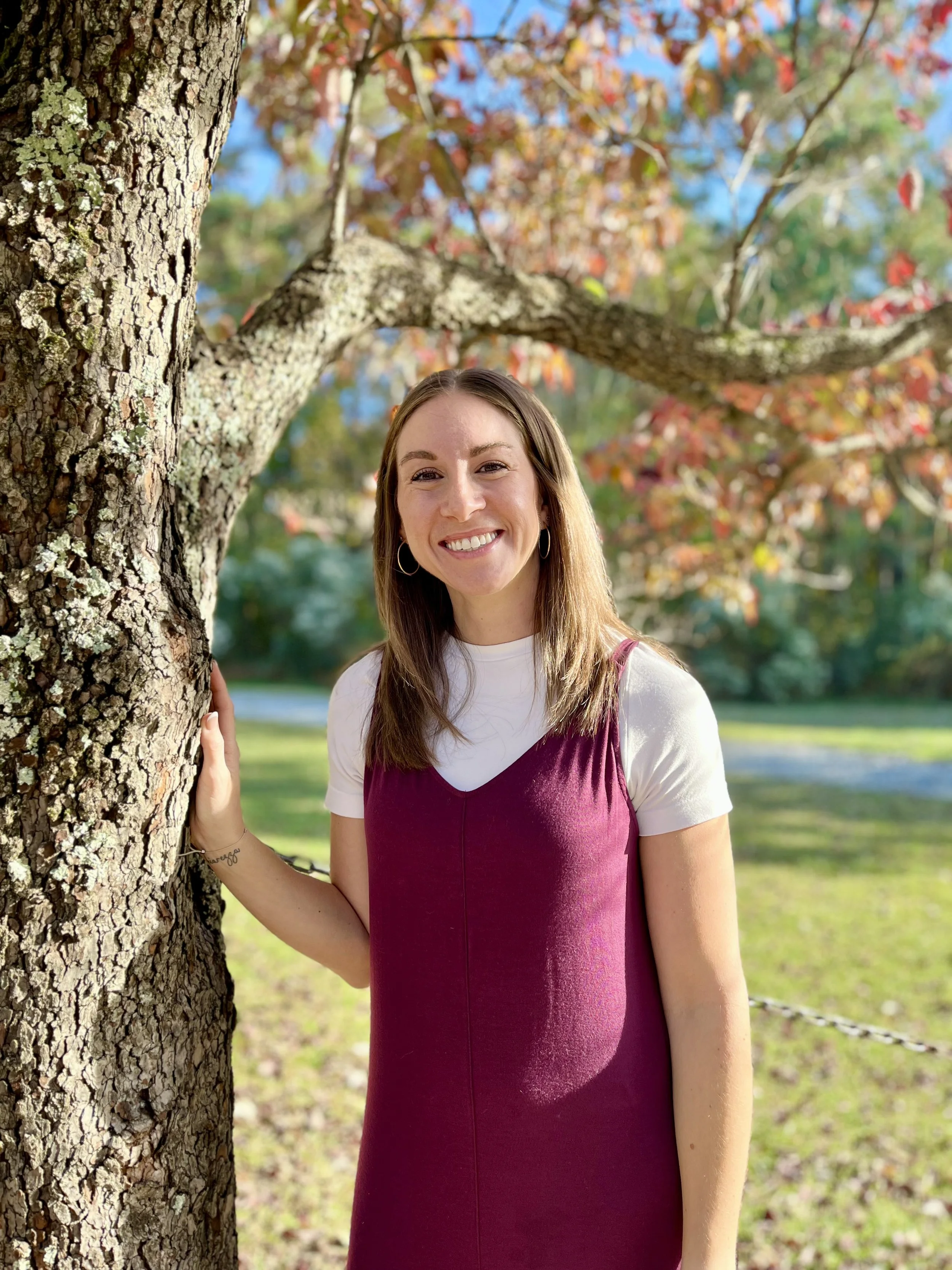 A young woman with long brown hair, smiling, standing outdoors near a tree with autumn-colored leaves, wearing a white t-shirt and a sleeveless maroon dress.