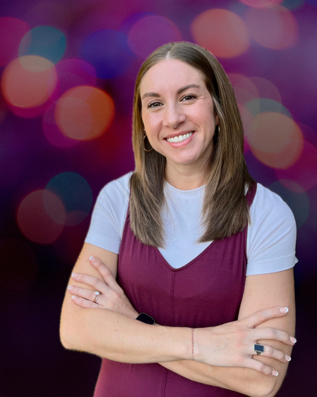 A smiling young woman with brown hair, wearing a white t-shirt layered under a maroon sleeveless dress, standing with arms crossed against a colorful bokeh background.