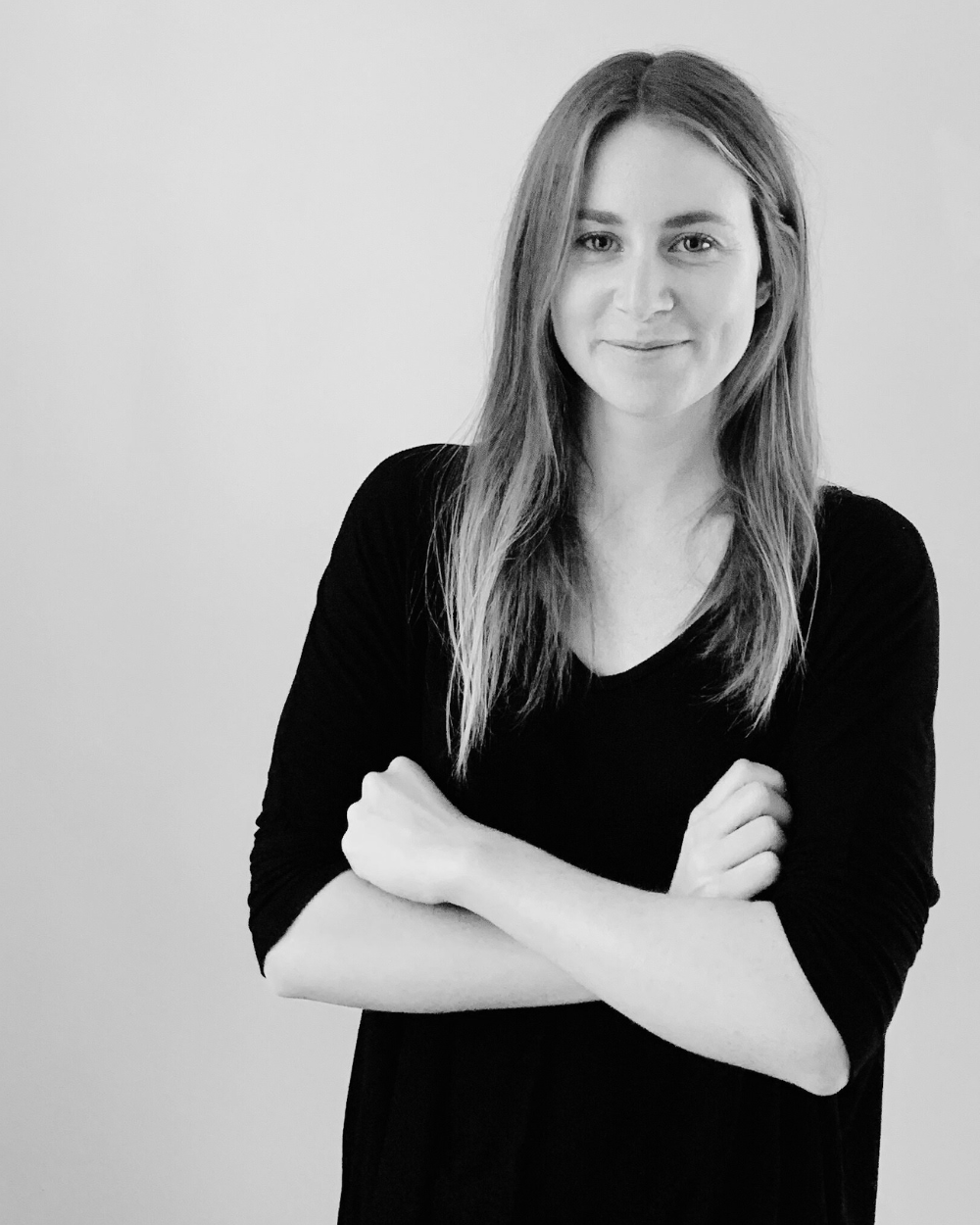 A woman with long hair crossing her arms, smiling, wearing a black top, standing against a plain background.