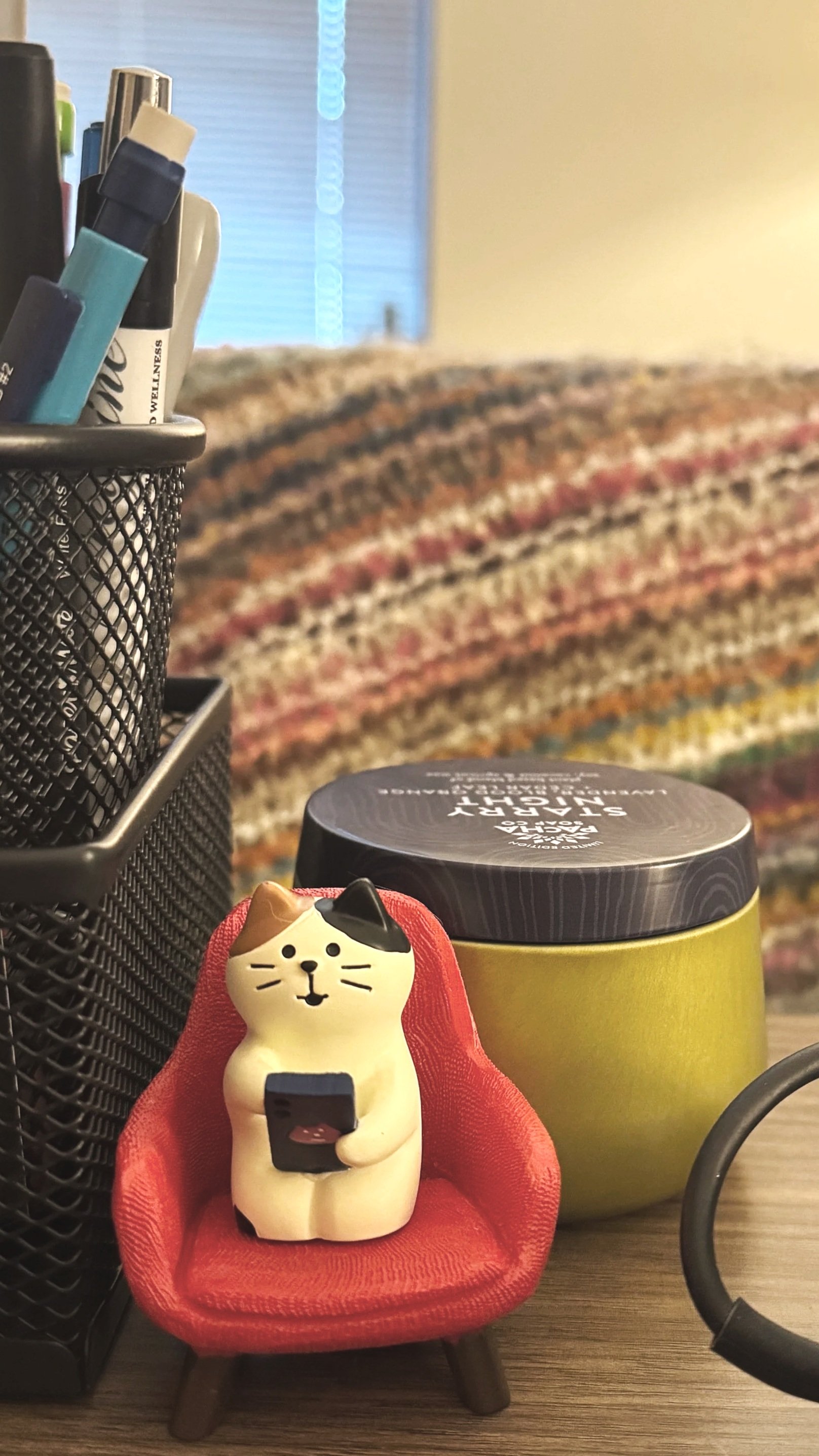 A decorative cat figurine sitting on a miniature red chair, holding a smartphone, next to a green container with a black lid, on a desk with a black pen holder and a colorful blanket in the background.