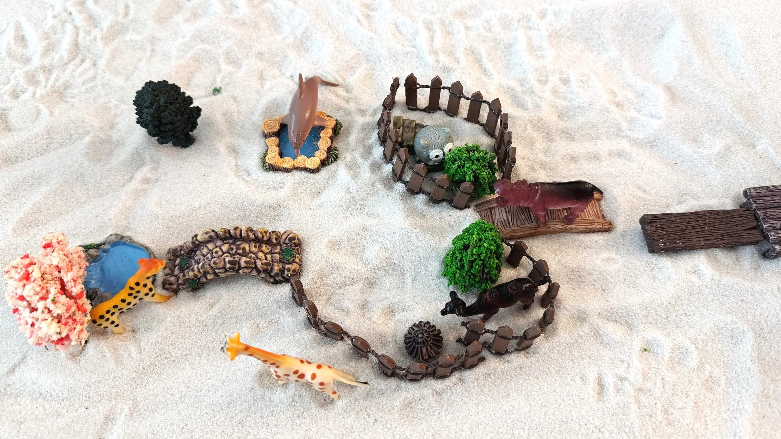 Toy animals including a giraffe, zebra, hippopotamus, crocodile, and various others arranged on sandy surface with miniature fences and trees.