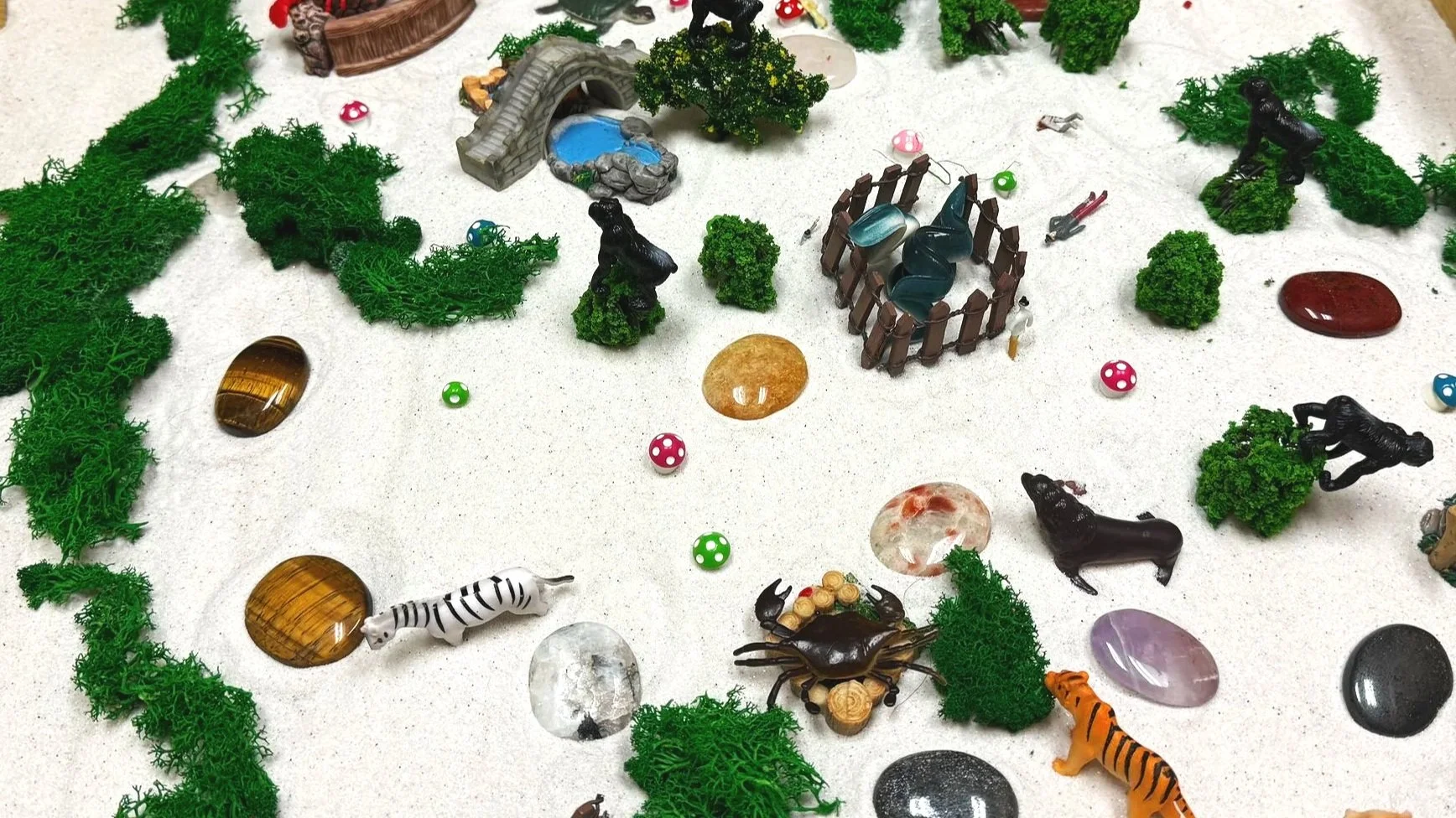 Miniature zoo scene with toy animals, rocks, trees, and decorative objects on a sandy surface.