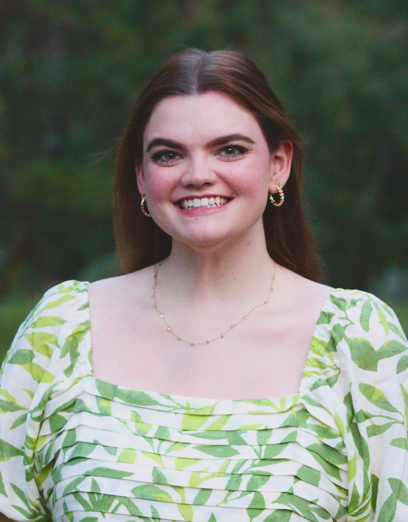 A young woman with long brown hair, blue eyes, and light skin smiling outdoors, wearing a green and white leaf-patterned dress, gold hoop earrings, and a delicate gold necklace, with a blurred green background.