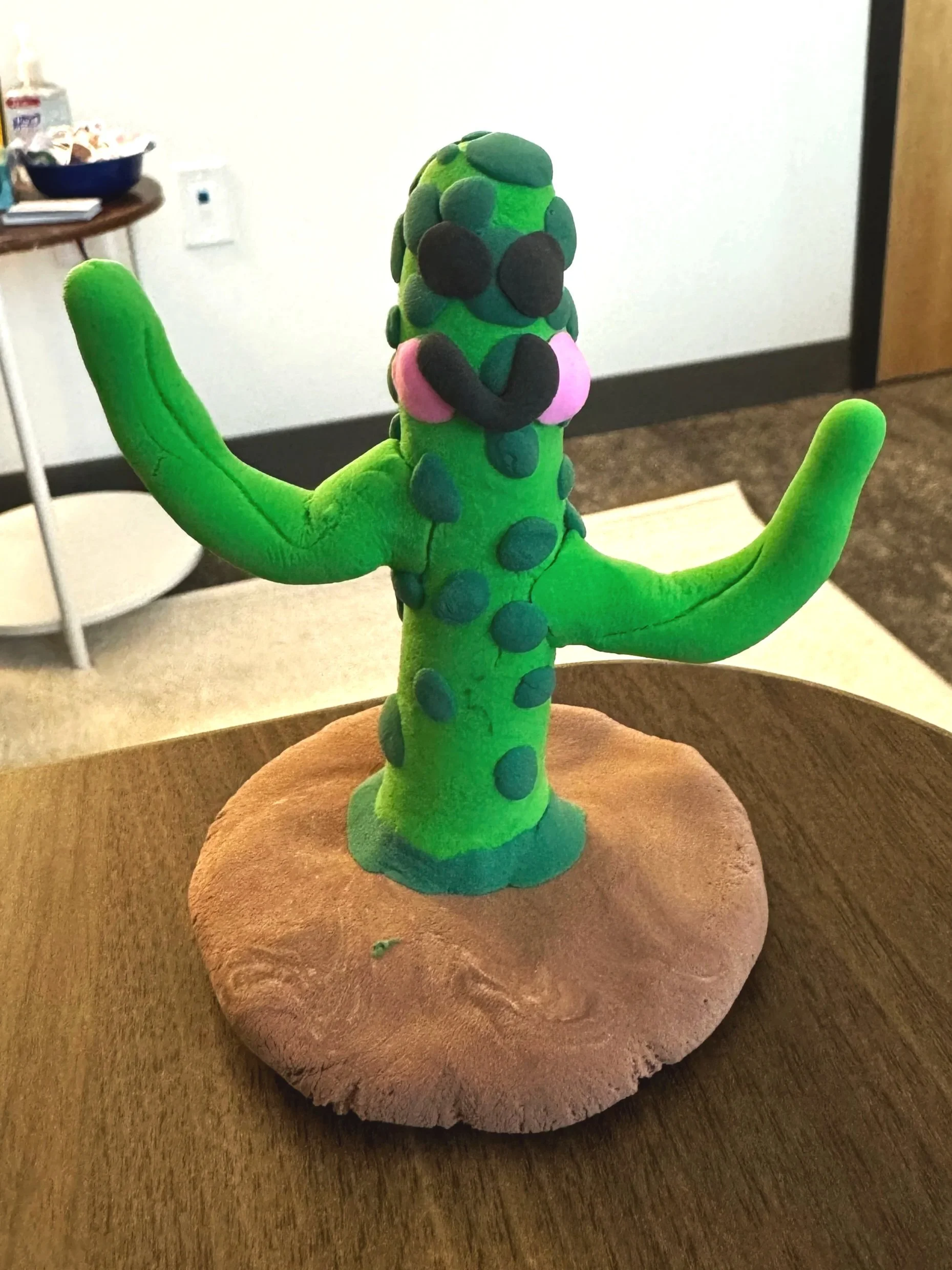 Handmade cactus sculpture with a smiling face, made of green and black modeling clay, displayed on a round base on a wooden table.