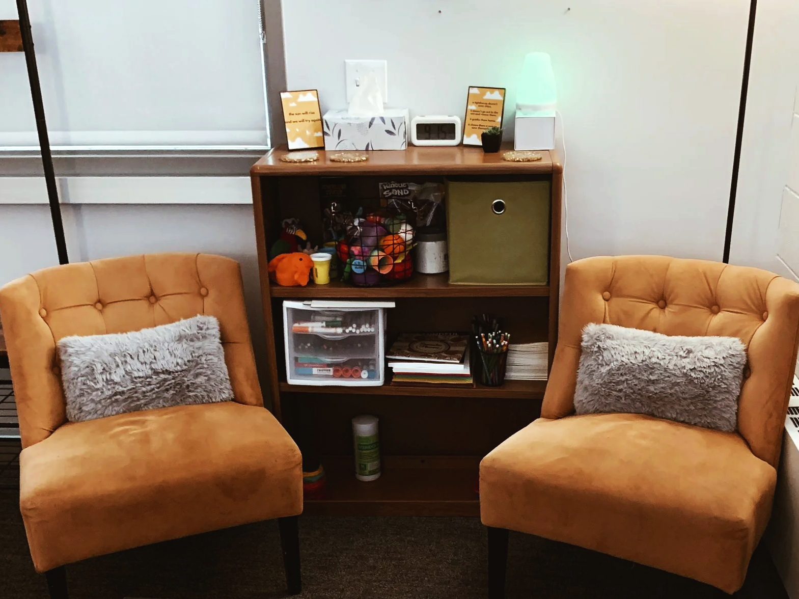 Two tan upholstered armchairs with gray furry pillows sit on a dark carpeted floor on either side of a wooden shelf. On top of the shelf are framed quotes, a digital clock, a lamp, tissue box, and small decorative items. The shelf contains toys, art supplies, books, and storage boxes.