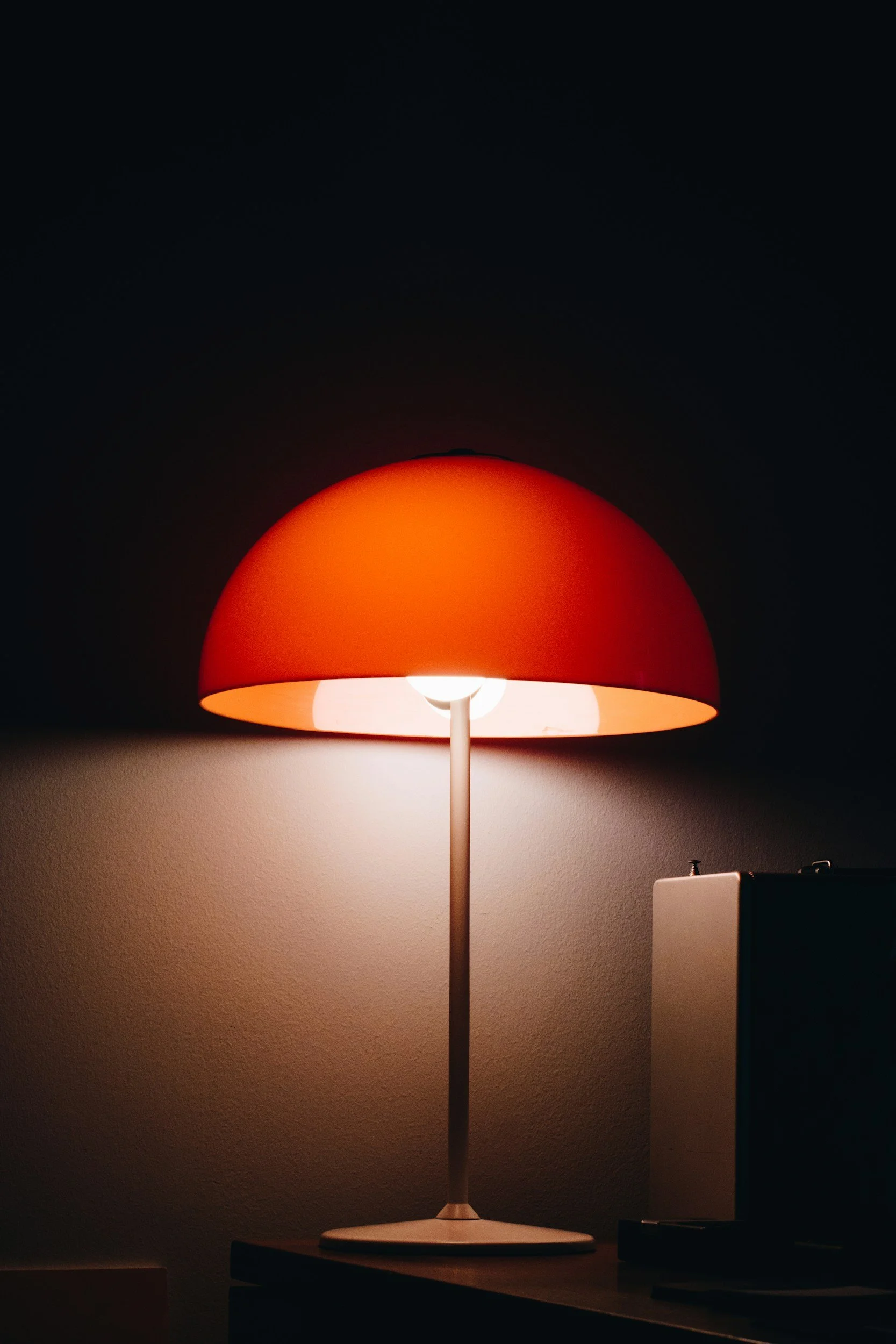 A modern table lamp with an orange lampshade emits light on a wall in a dark room.