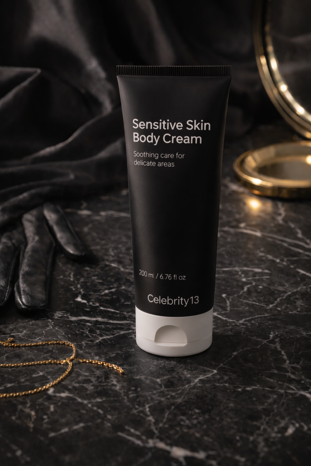 A black tube of sensitive skin body cream on a dark marble surface, surrounded by black leather gloves, a gold chain, and a gold-rimmed mirror in the background.