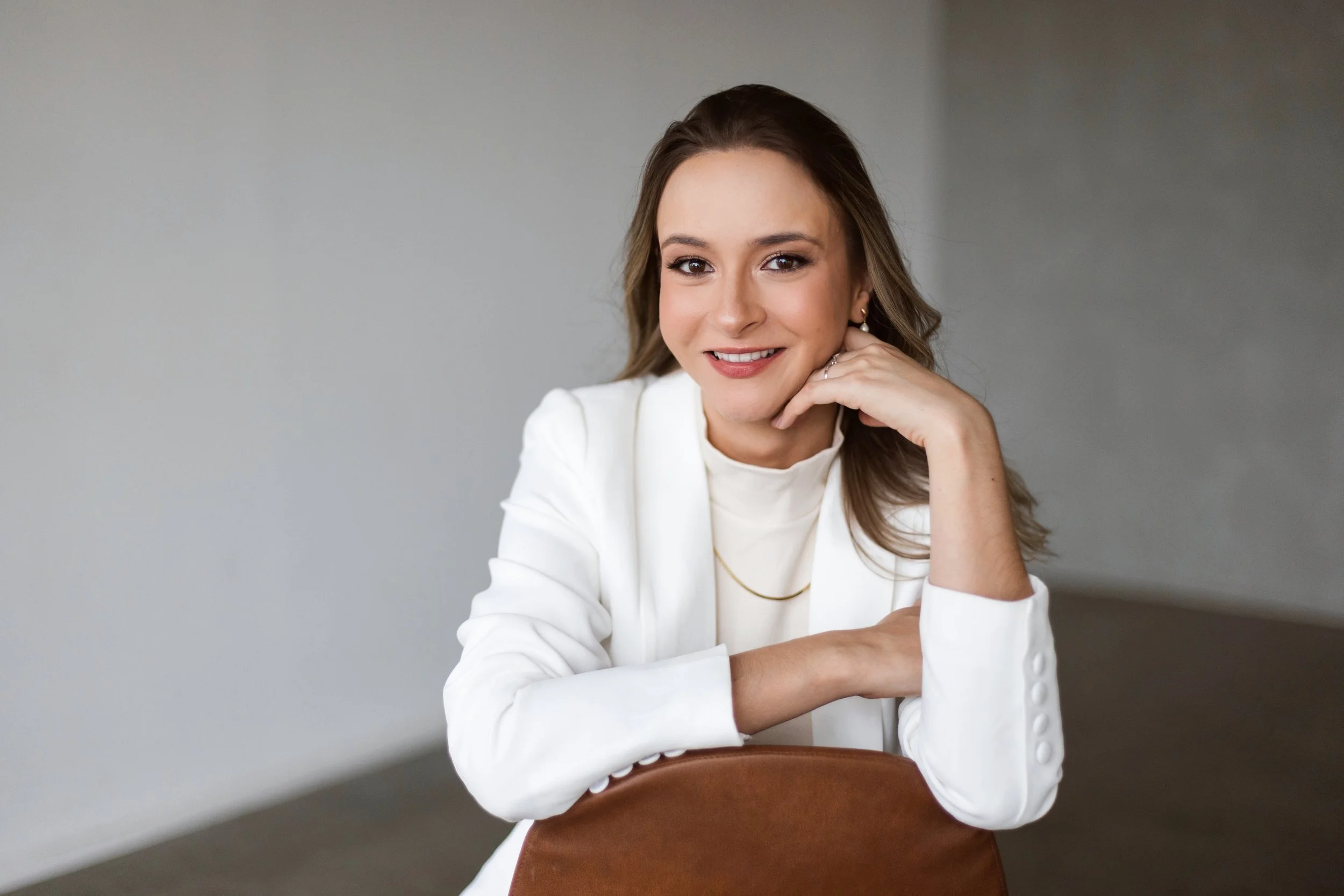 Mulher sorridente com cabelo castanho claro, usando blusa branca e blazer branco, sentada em uma cadeira, apoiando o queixo na mão, em um ambiente de fundo neutro.