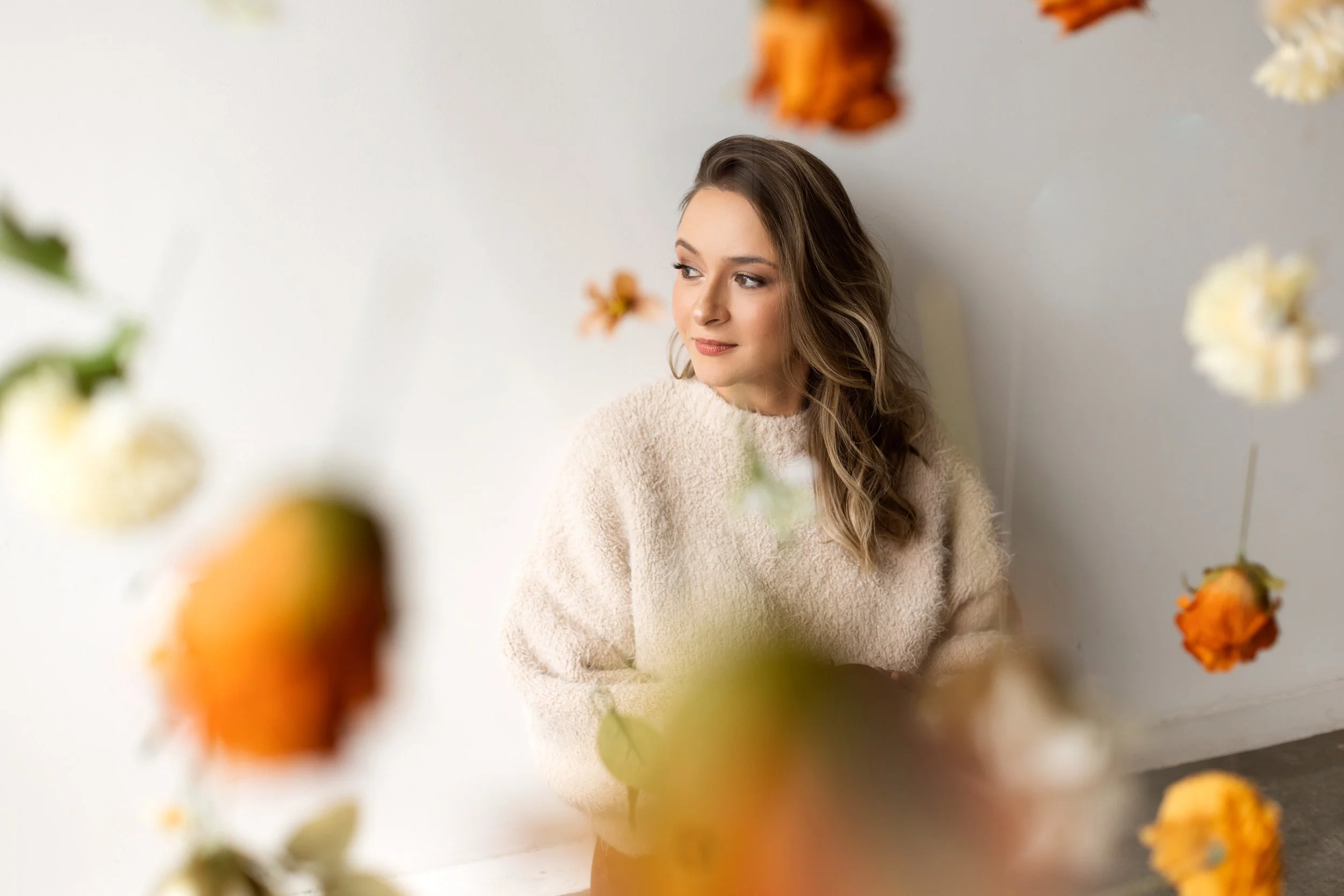 Mulher de cabelo castanho claro e ondulado, vestindo roupa de lã bege, olhando para o lado, com flores borradas na frente e ao redor, contra parede branca.