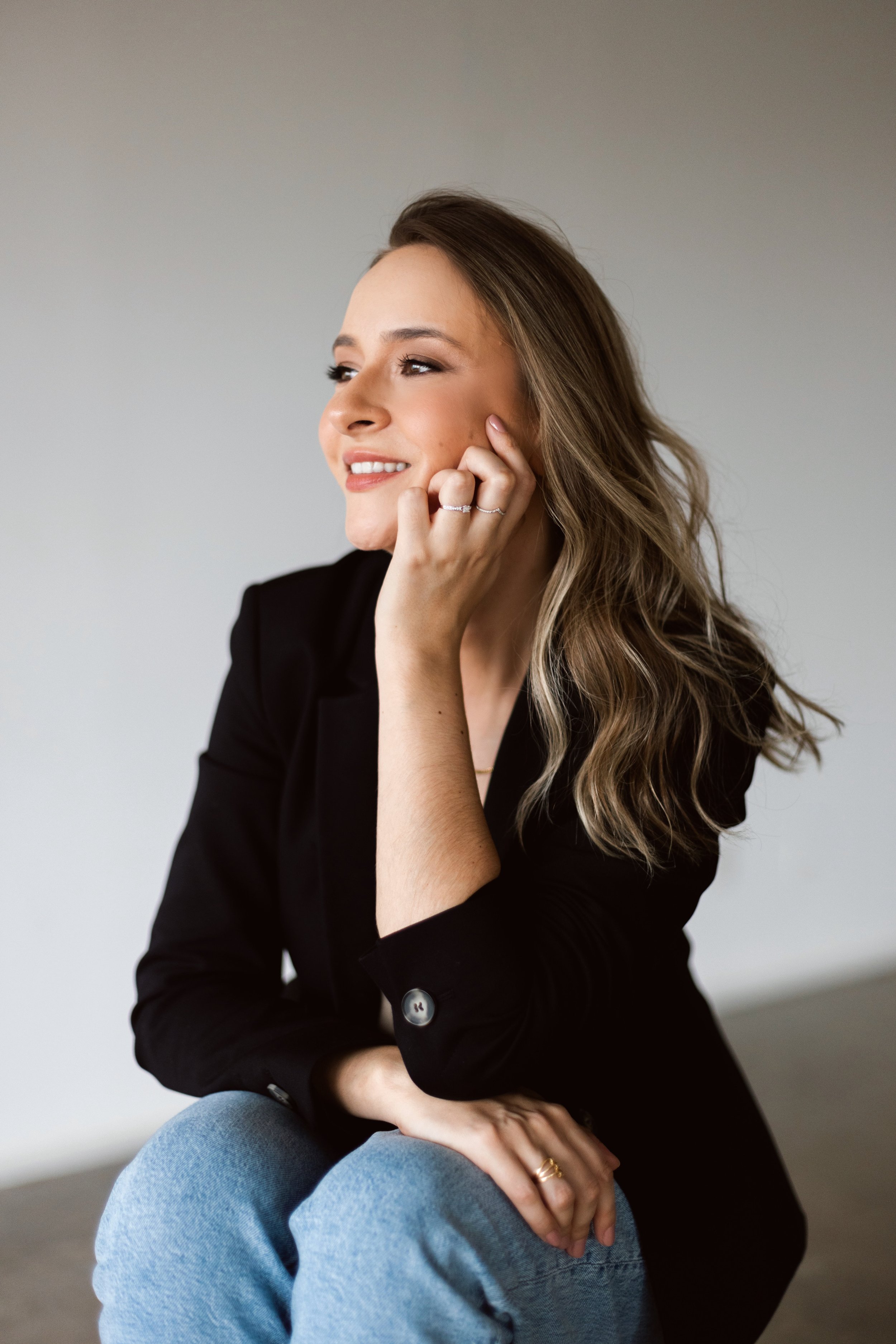 Mulher sorrindo, de cabelo loiro com cachos soltos, usando blazer preto e jeans claros, sentada com a mão no rosto, olhando para o lado.