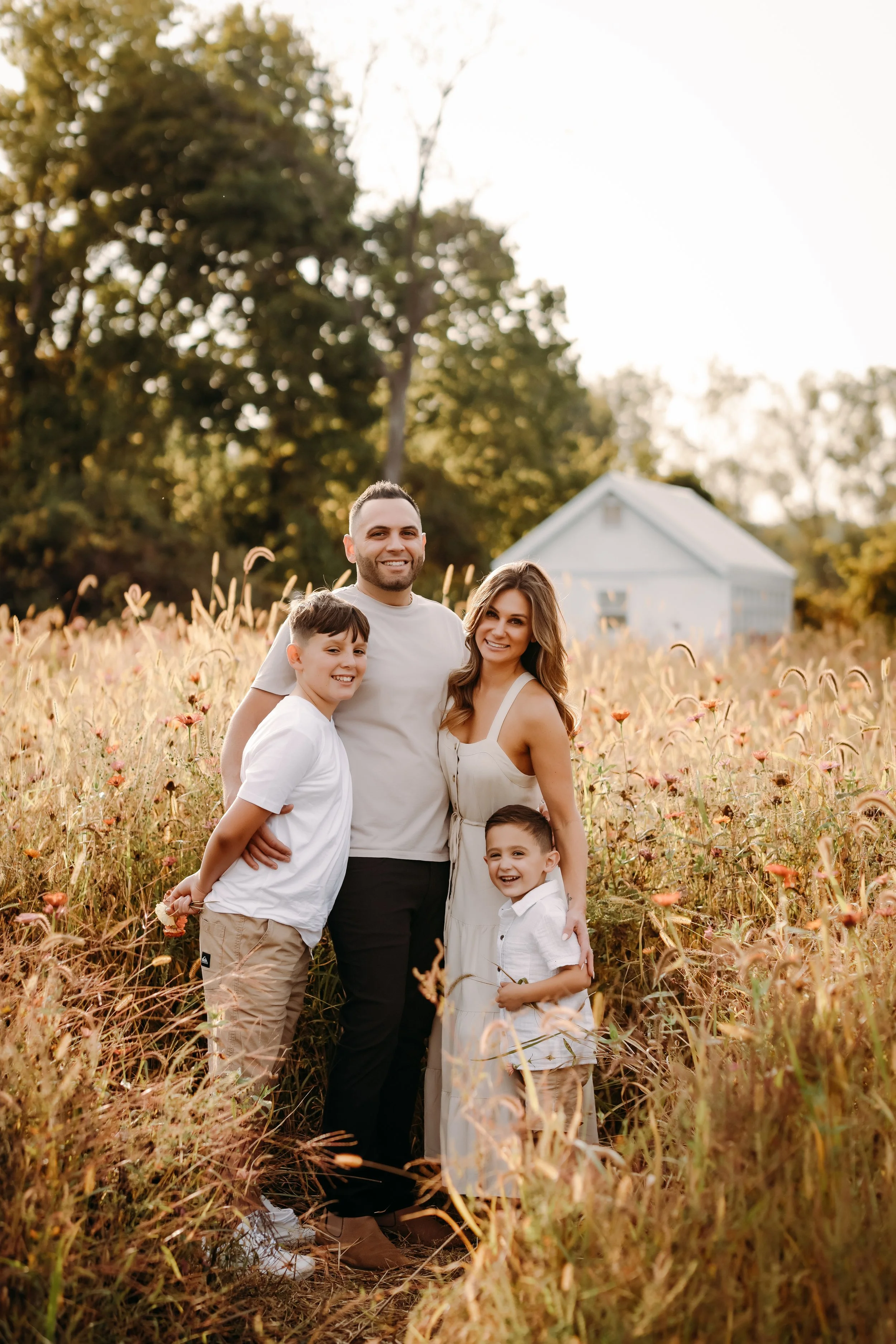 A family of five standing in a field of tall grass and flowers, smiling at the camera with a white house and trees in the background during golden hour.