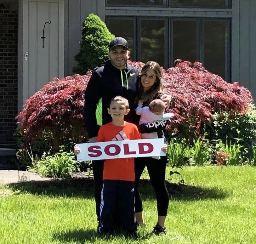 Family of four celebrating sale of their house outside in front of a bush with red leaves, holding a 'SOLD' sign.
