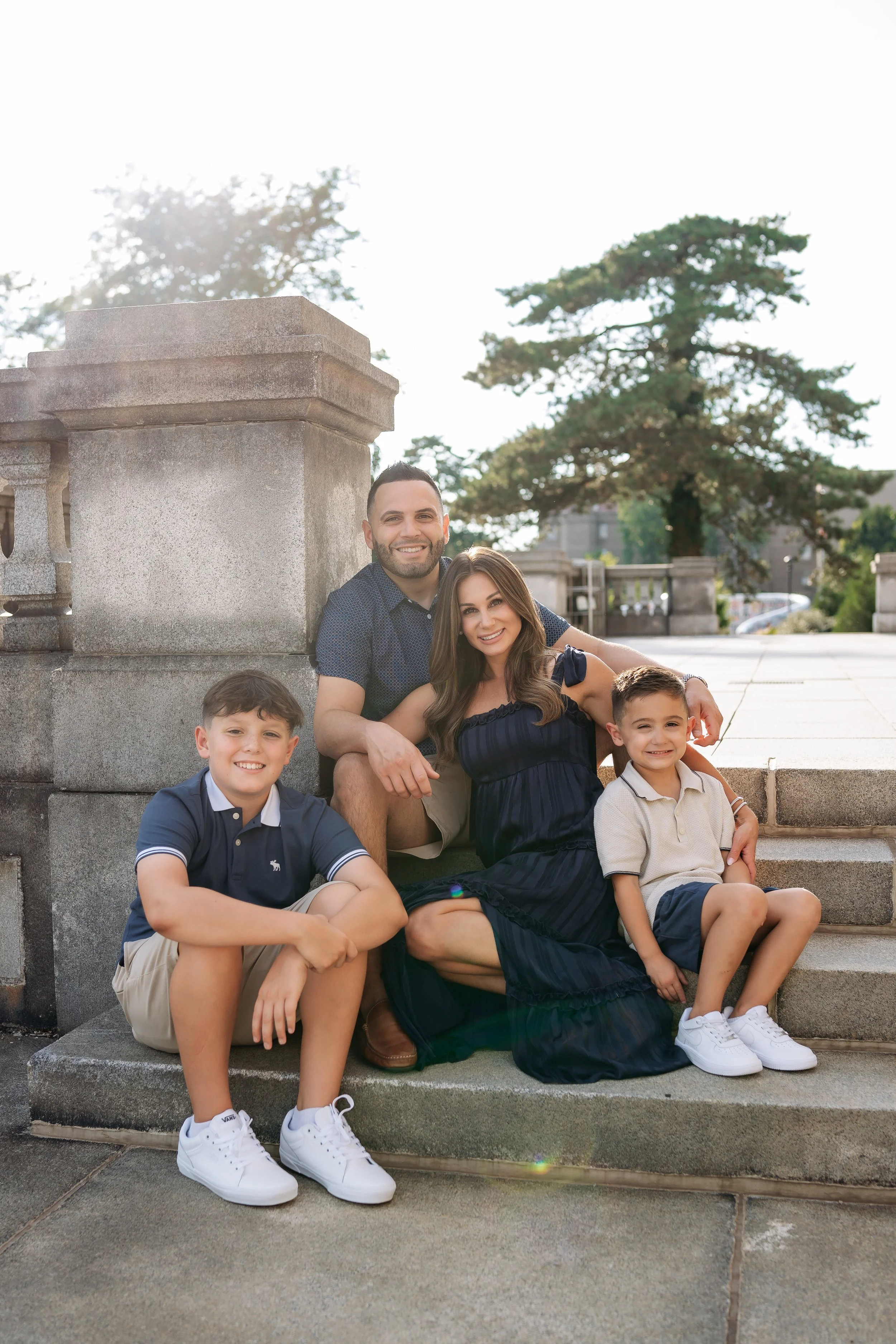A family of five sitting on steps outdoors in front of a large tree and a stone railing, smiling at the camera during daytime.